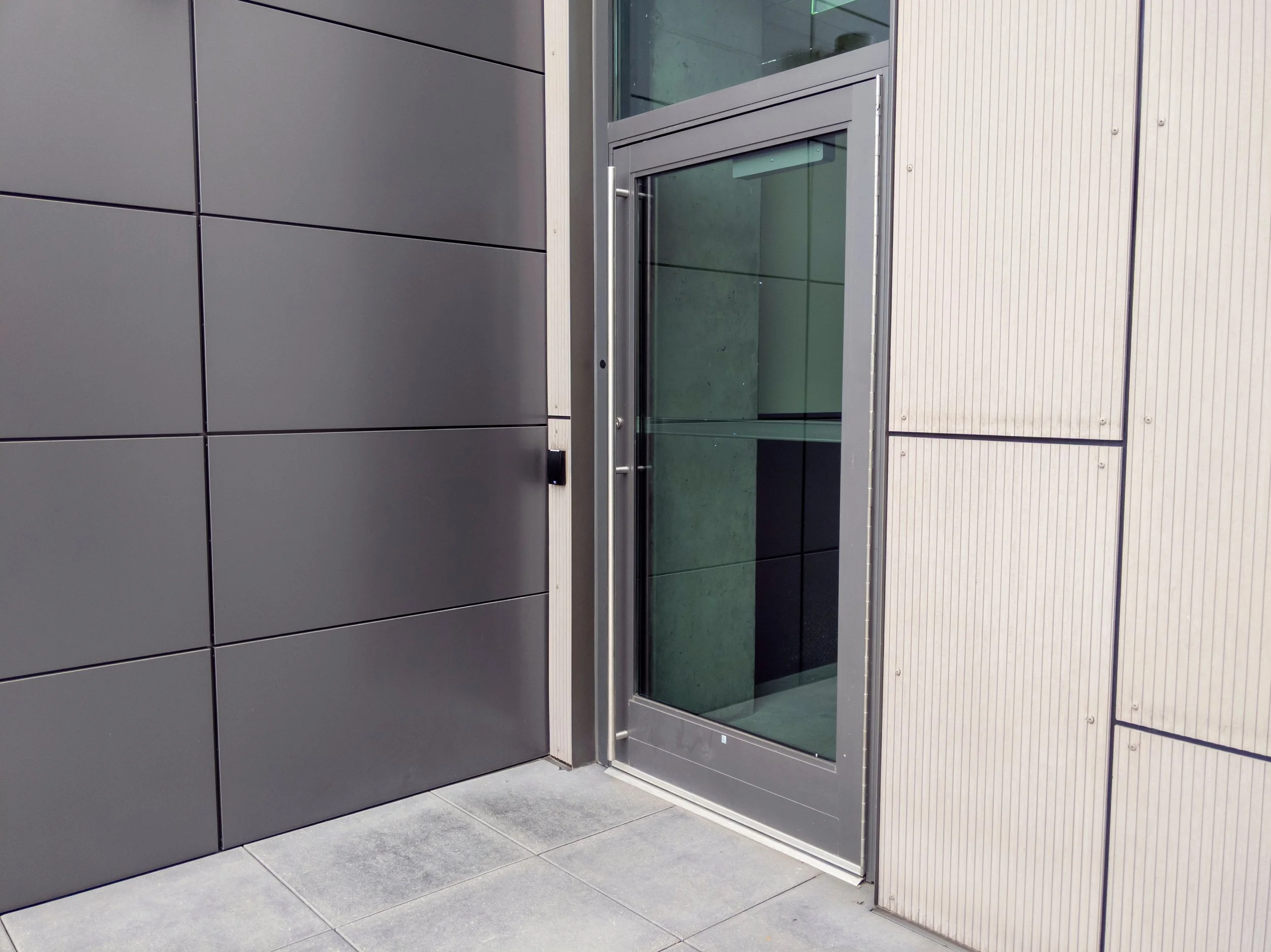 Modern building entrance with dark gray panel walls, a glass door with an aluminium frame, and a light-colored textured wall panel on the right side.