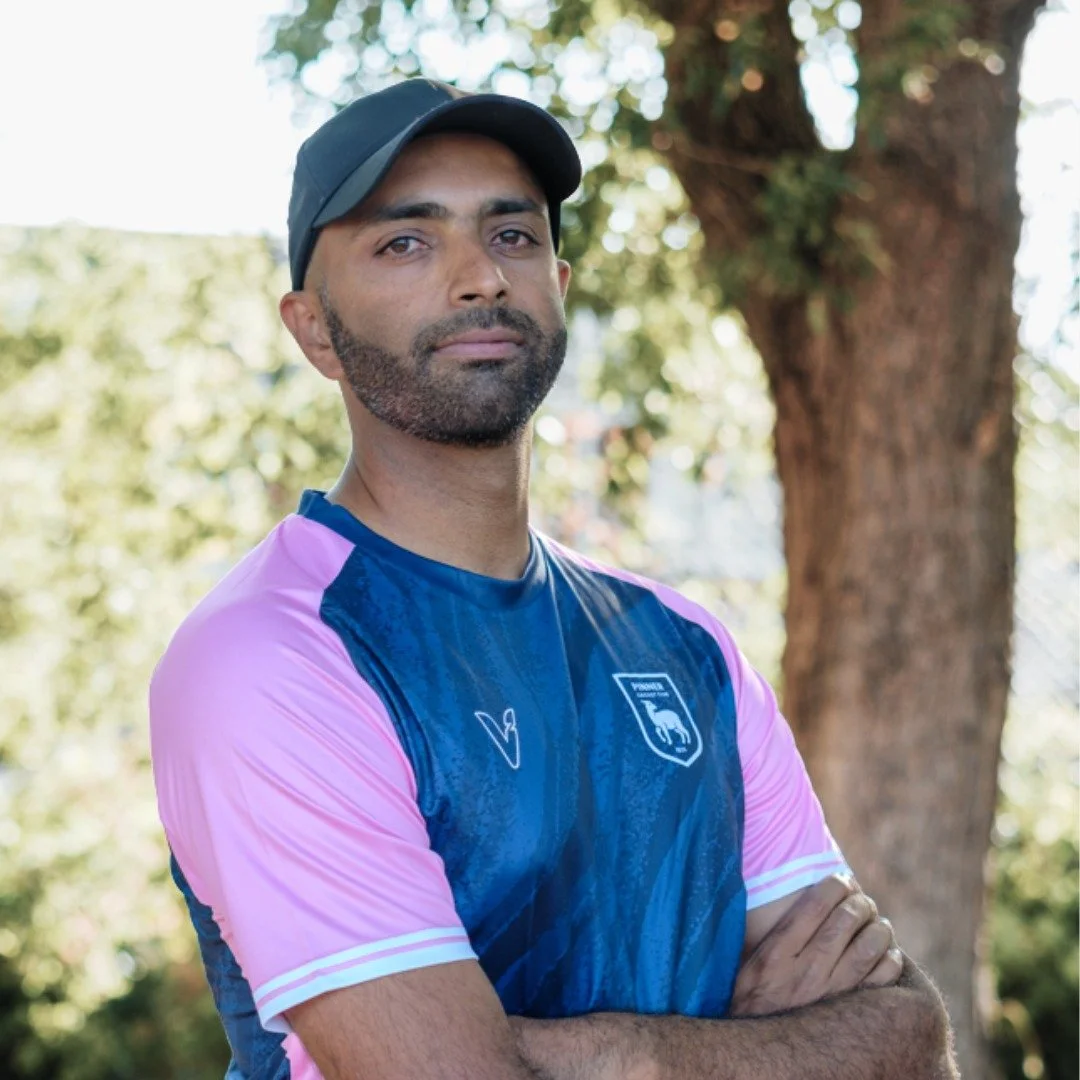 A man with medium skin tone, dark hair, and a beard, wearing a black cap and a sports jersey with pink sleeves and a blue front, standing outdoors with arms crossed in front of a tree, with green foliage in the background.