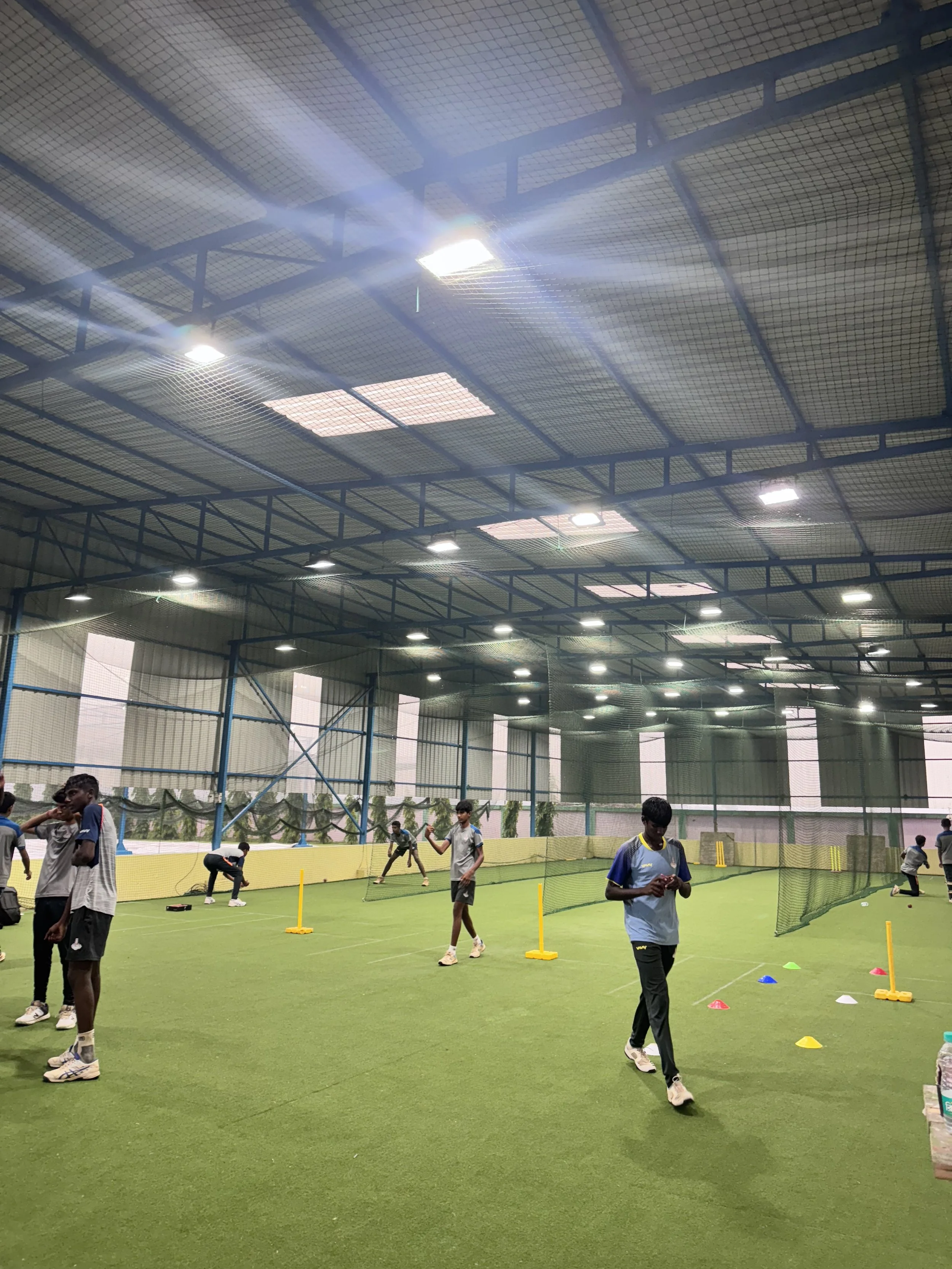 Indoor sports facility with children practicing cricket skills, using cones and poles for drills, under a high ceiling with lights and netting.
