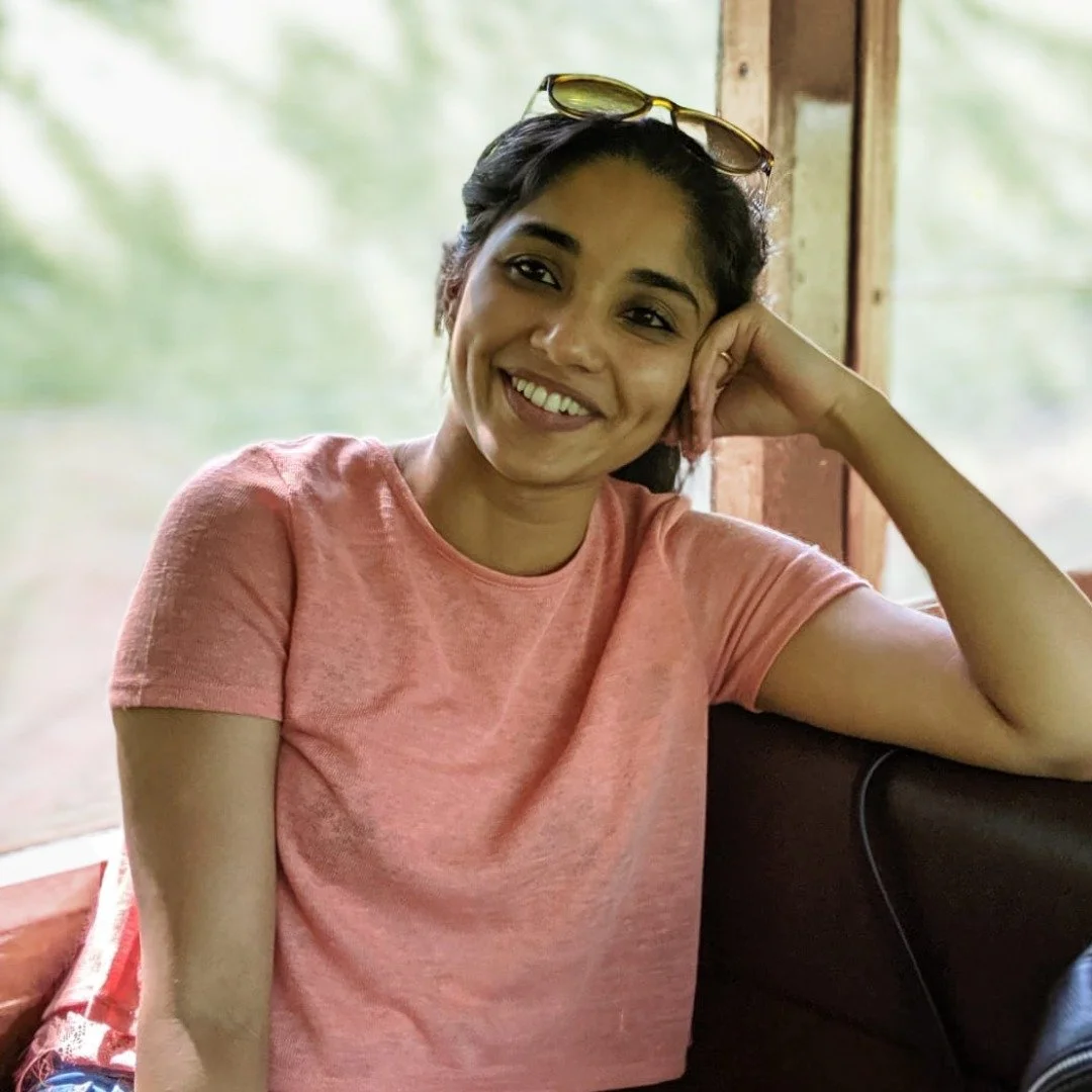 A woman with dark hair, wearing a pink t-shirt and sunglasses on her head, sitting indoors near a window with a relaxed posture and smiling.