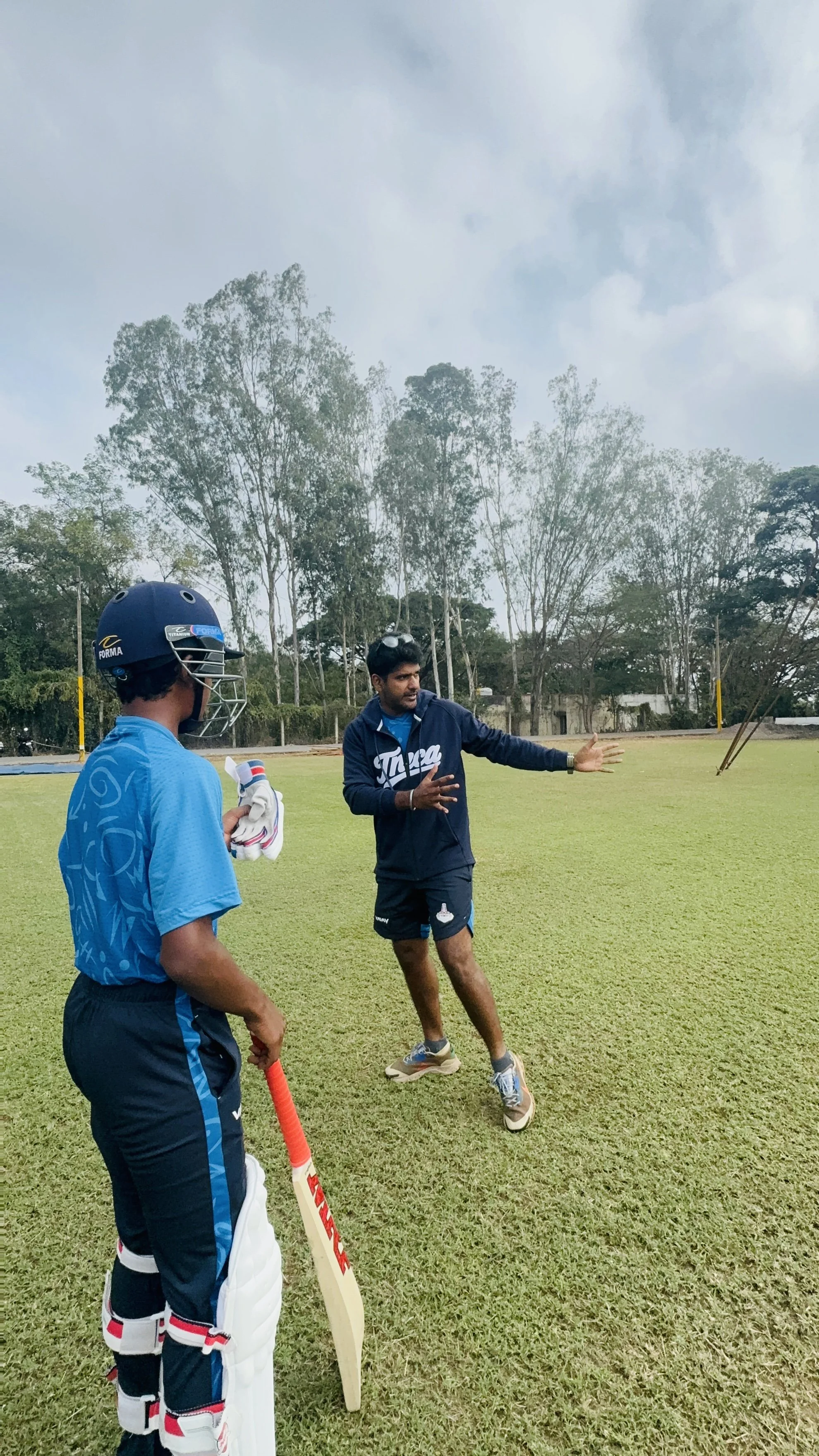 Two young cricket players are on a grassy field, standing and listening to their coach who is demonstrating cricket techniques. One player is in a batting protective gear and helmet, holding a cricket bat, while the coach, dressed in casual athletic clothing, is gesturing with his hand.