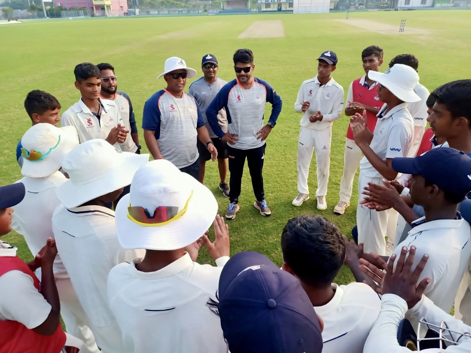 Cricket team gathered in a circle on a field, possibly during a team meeting or coaching session, with players wearing white uniforms and wide-brimmed hats and some individuals in casual attire, with a cricket pitch visible in the background.