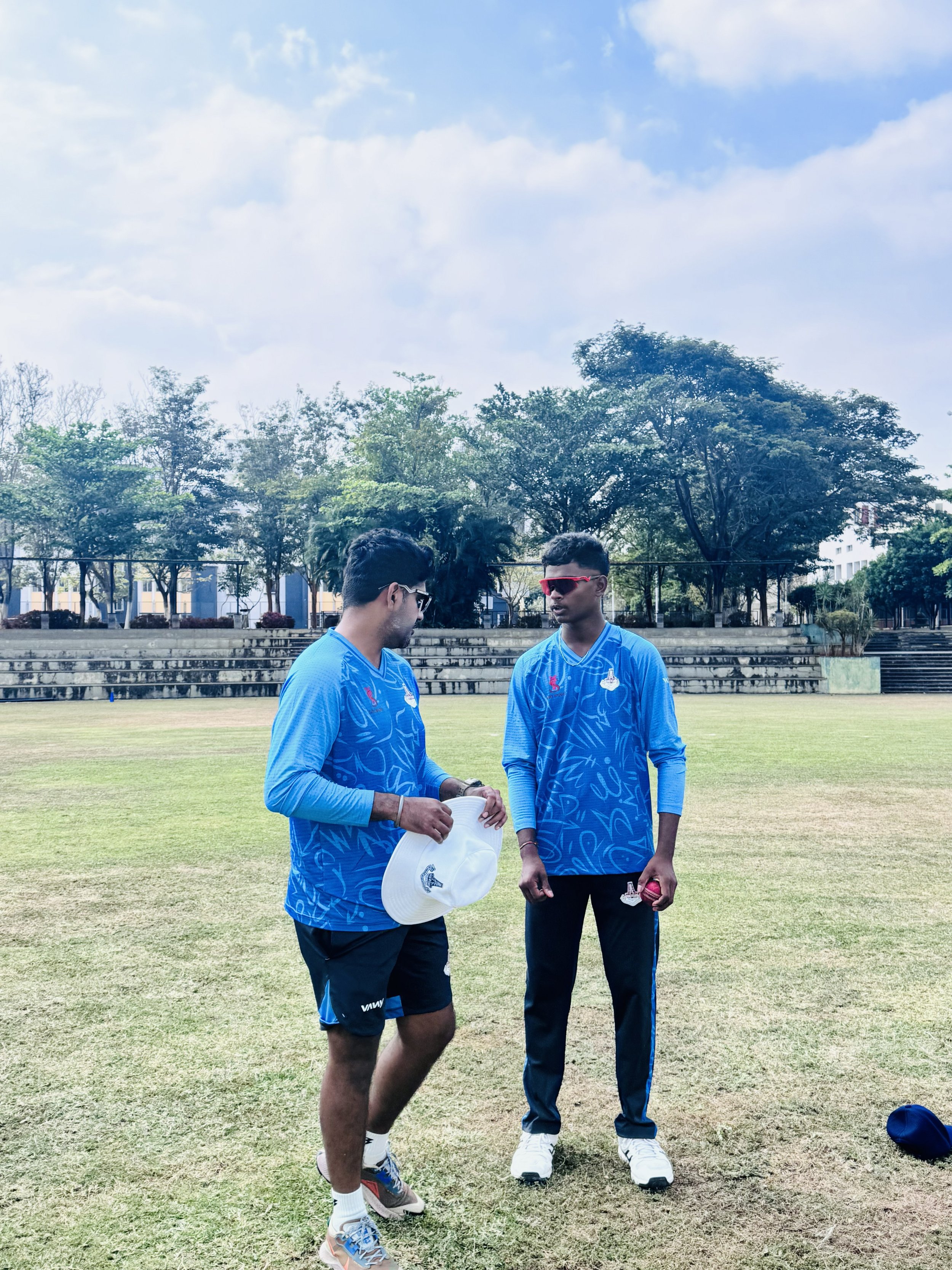 Two young male cricket players wearing blue jerseys and black shorts standing on a grass field, engaged in conversation, with trees and a sports stadium in the background.