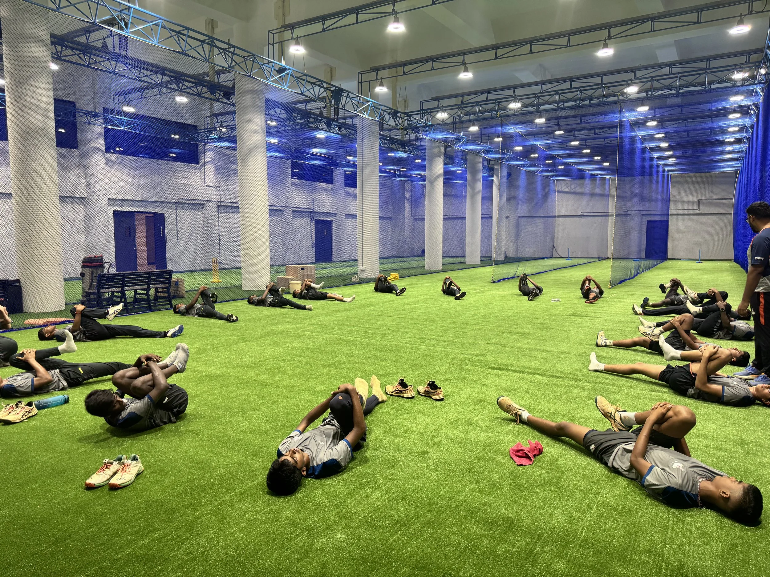 Group of people in sports attire practicing yoga or stretching on artificial turf inside an indoor sports facility with blue netting and high ceilings.