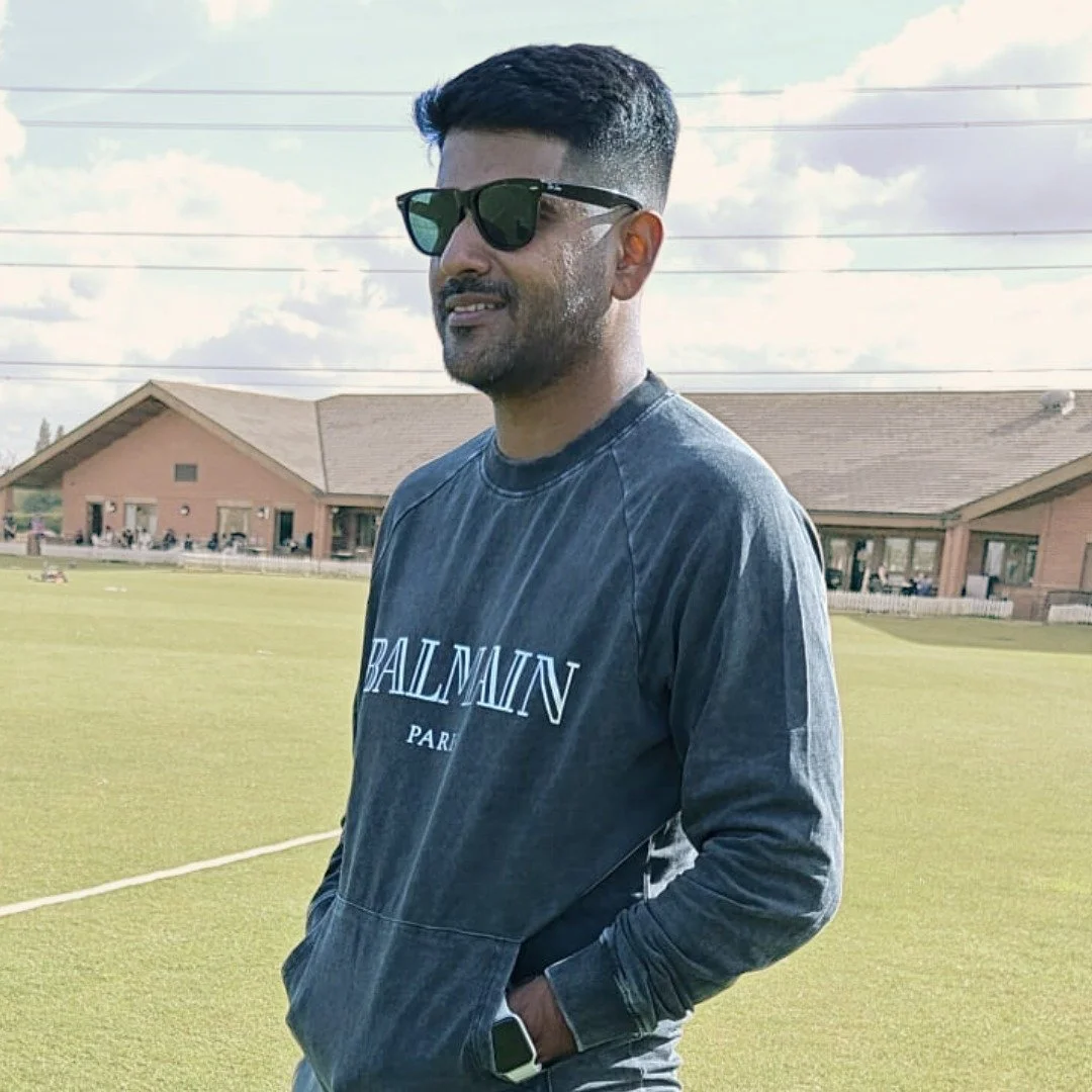 A man with short dark hair and sunglasses standing outdoors on a sports field, wearing a black Balmain Paris sweatshirt and a smartwatch, with buildings and a partly cloudy sky in the background.