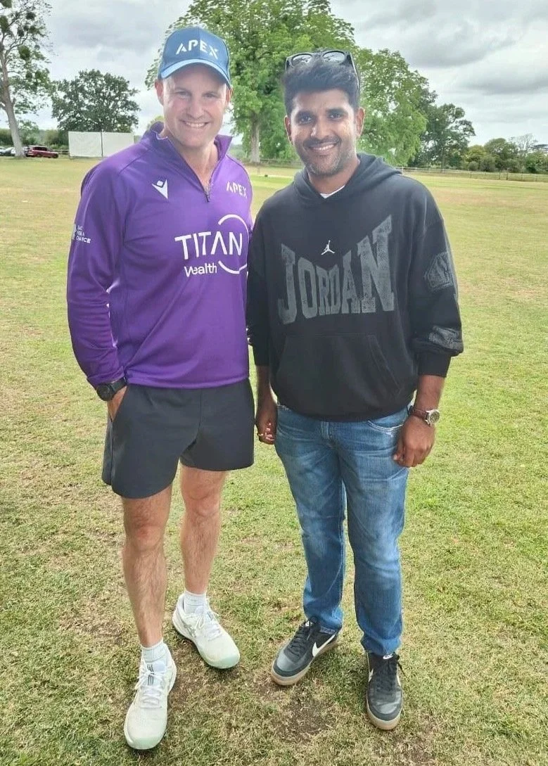 Two men standing on a grassy field with trees in the background, smiling at the camera. One is wearing a purple jacket, black shorts, and white sneakers, while the other is wearing a black hoodie, blue jeans, and gray sneakers.