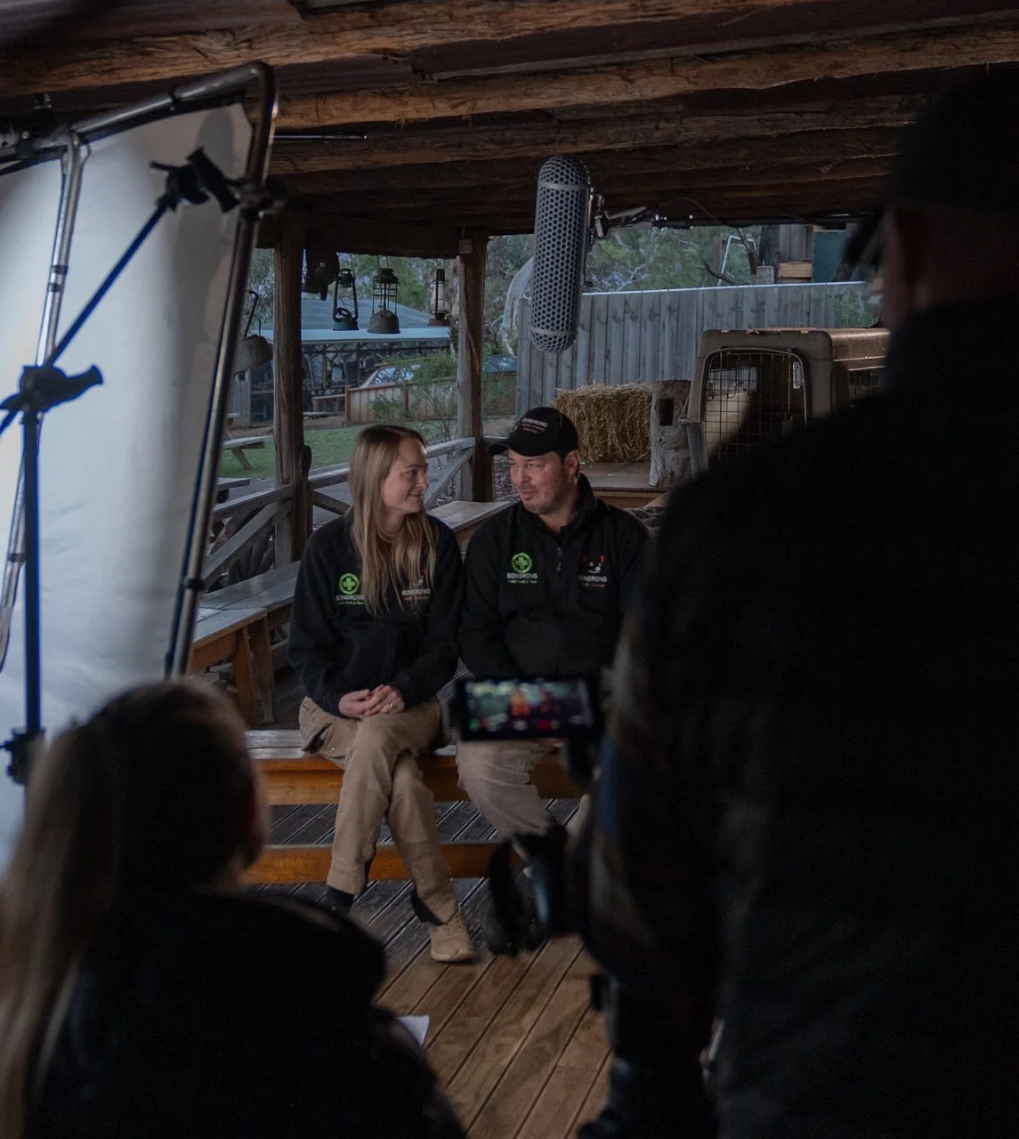 Behind the scenes photo of Greg and Melissa Irons doing an interview for the docuseries the Animal Sanctuary.  Sitting in a rustic wooden hut