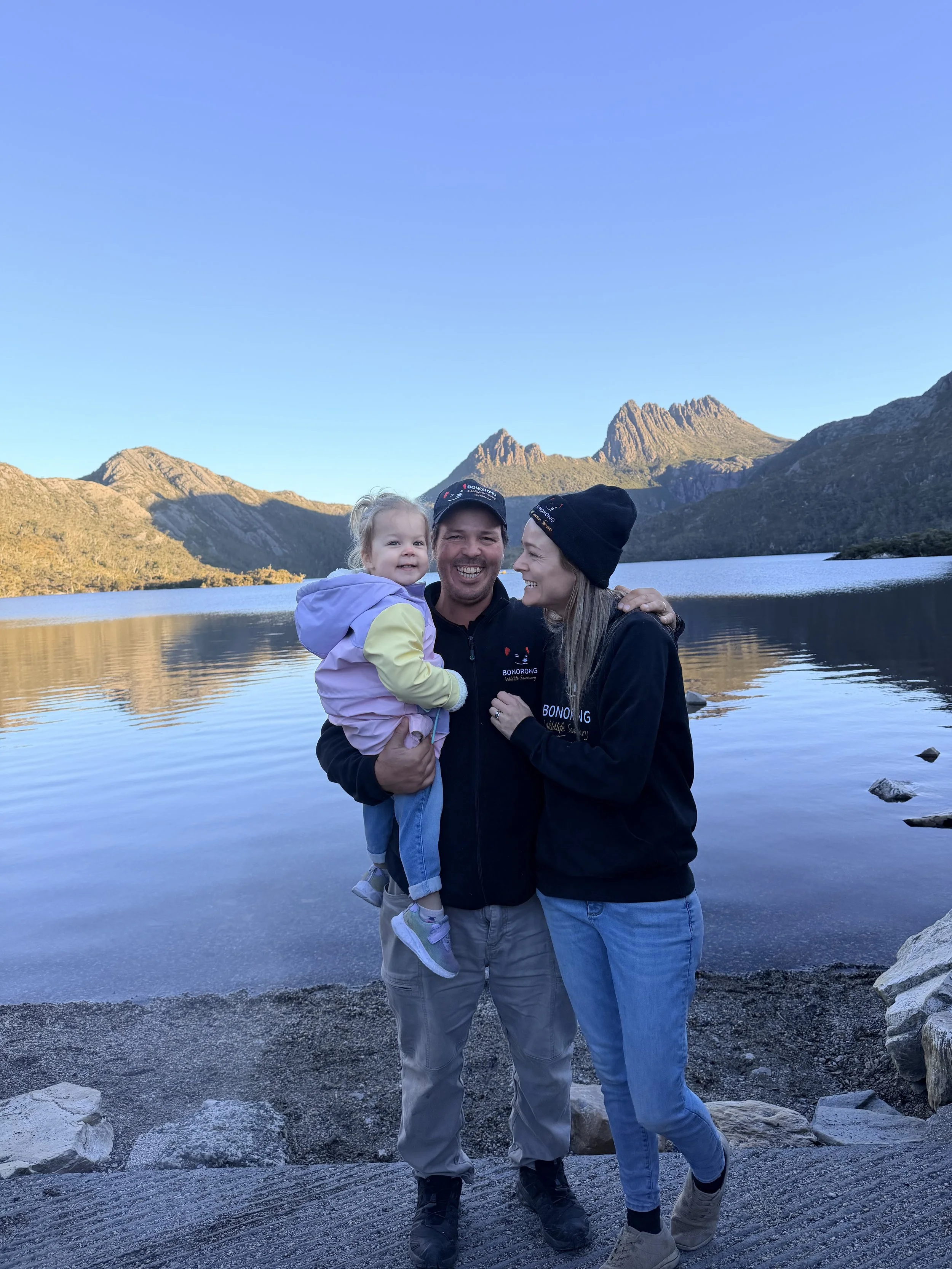 Greg Irons, Melissa Irons, and Lara Irons, standing together by Dove Lake with Cradle Mountain in the background, smiling and enjoying the outdoors during daytime.