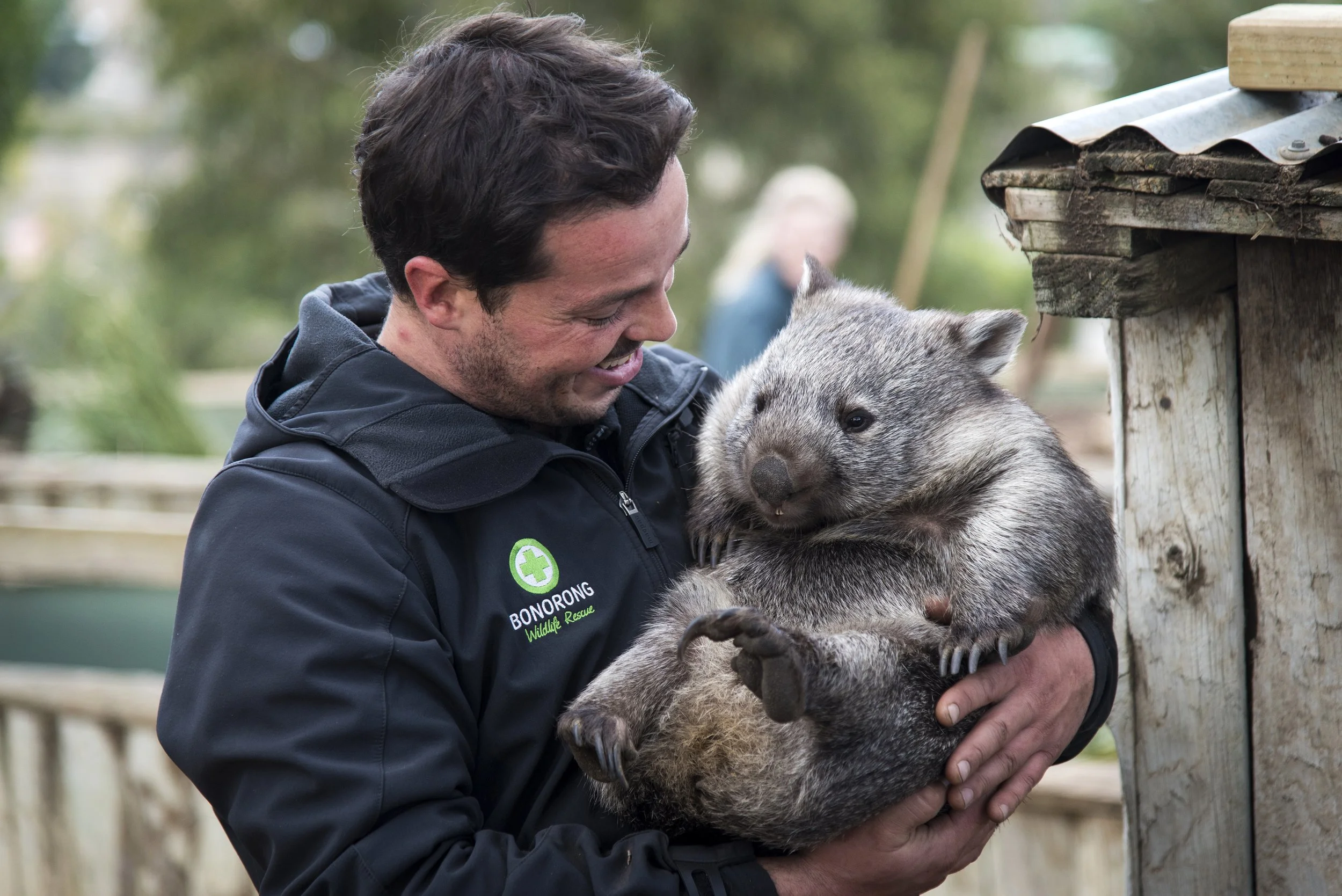 Greg Irons holding a baby wombat outdoors, smiling and looking at the wombat, with trees in the background.
