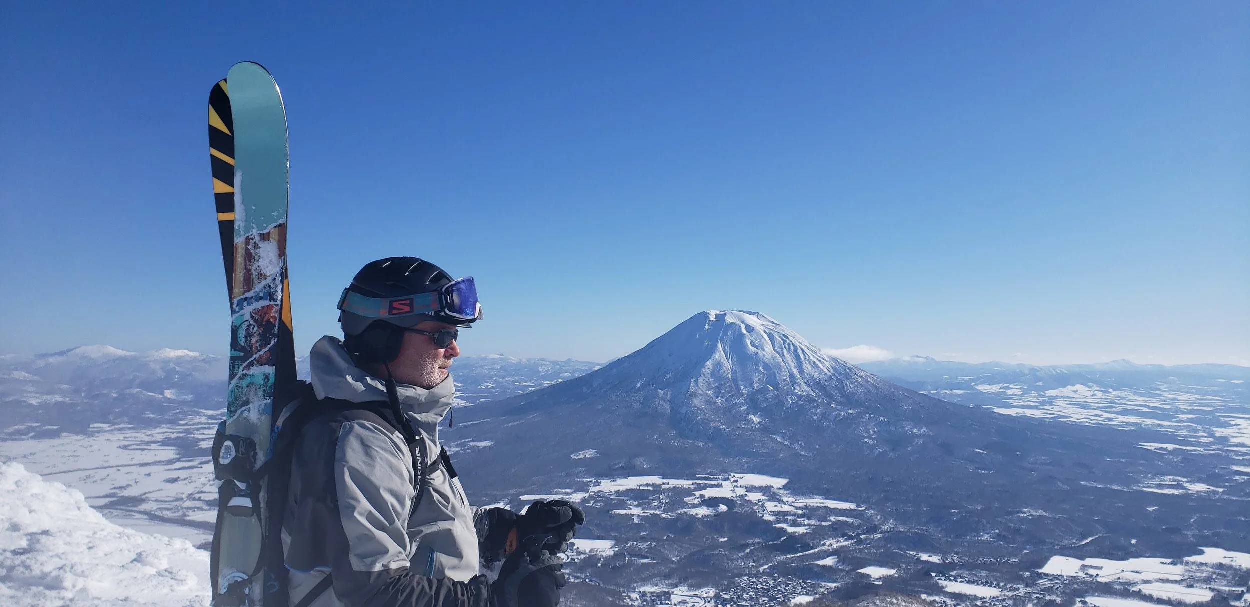 A person in winter gear, wearing a helmet and sunglasses, stands on a snowy mountain with skis on their back, overlooking a snow-covered landscape with a volcano and plains in the distance. Mt. Yotei, Hokkaido, Japan.