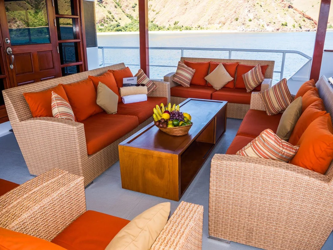 A cozy outdoor seating area on a boat with wicker sofas and orange cushions, overlooking a body of water with hills in the background.