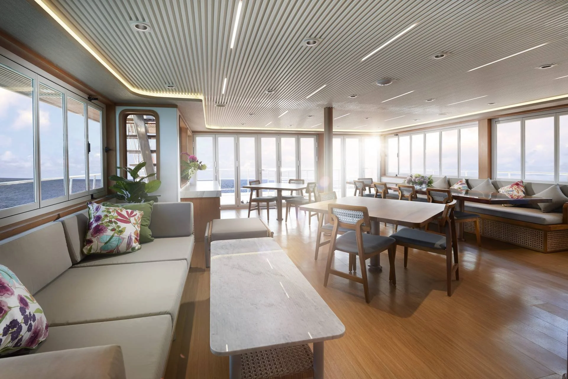 Bright indoor lounge area on a cruise ship with windows overlooking the ocean, featuring sofas, chairs, and decorative pillows.