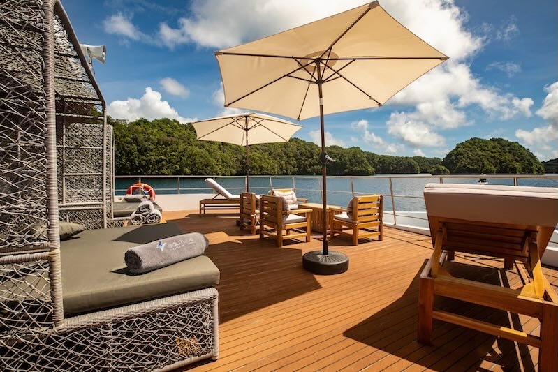 A large outdoor deck with beige umbrellas, wooden chairs, a cushioned lounge chair, and a view of water and trees in the background.