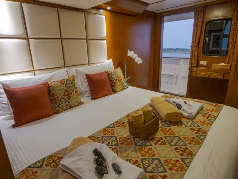 A hotel room with a large bed, patterned pillows, and towels. There is a sliding glass door leading outside with a view of water.