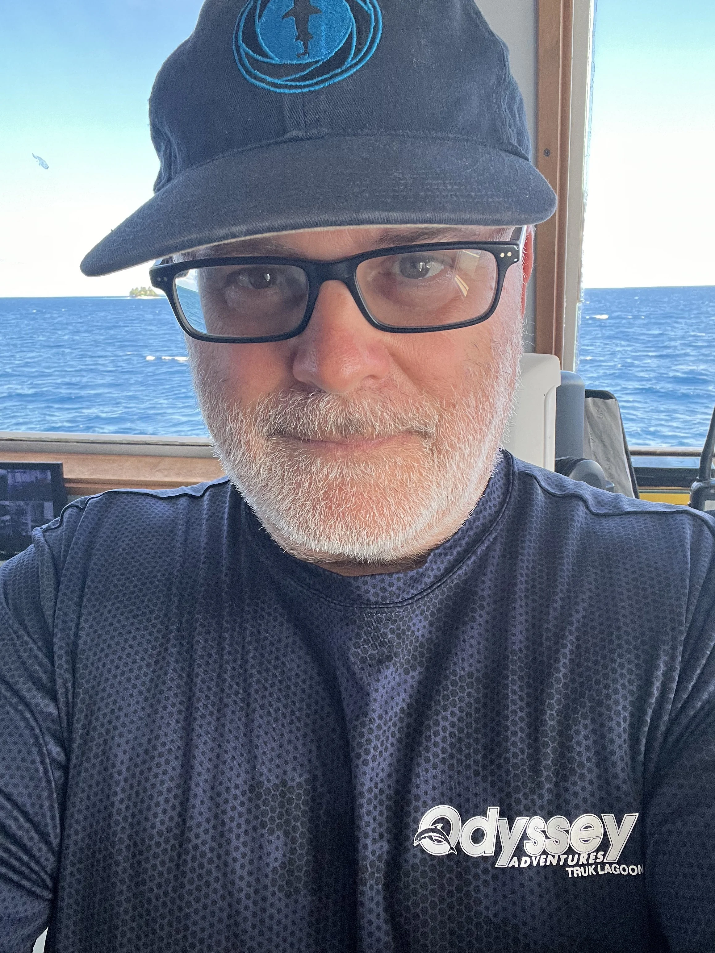 Close-up of a man wearing black glasses, a black cap with a blue logo, and a black t-shirt with the words 'Odyssey Adventures Truk Lagoon' printed on it. Behind him, there is a window showing the ocean and a small island in the distance.