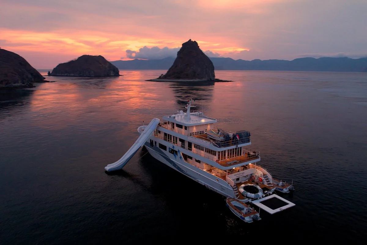 A large yacht with a water slide on the side, floating on calm water near rocky islands during a sunset.