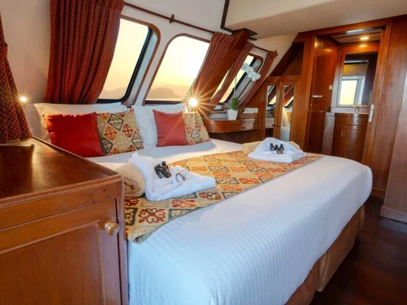 A cozy hotel room with a large bed, orange and patterned pillows, and a blanket, surrounded by wooden furniture and a window with curtains showing a sunset view.