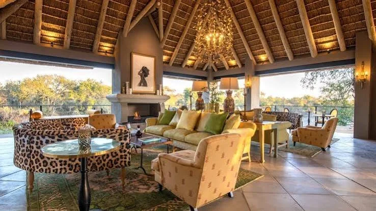 A spacious living room with a high, thatched ceiling and large windows. There is a yellow couch with green and beige pillows, a leopard print armchair, and a patterned armchair. A fireplace with artwork on the mantel is centered on the wall. Lamps and decorative vases are on the side tables, and a chandelier hangs from the ceiling. The room overlooks a balcony with trees and a scenic view.