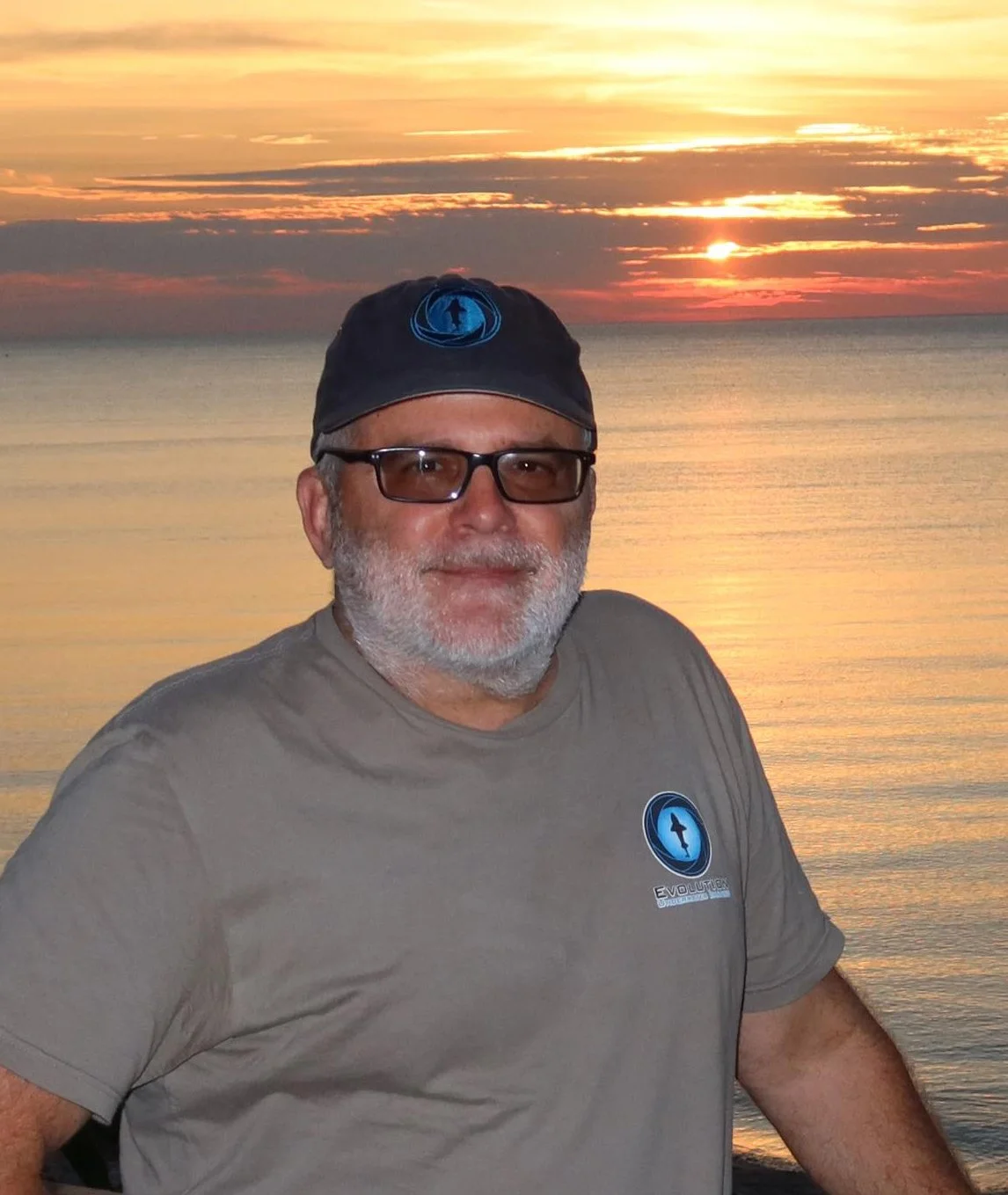 A man with a beard and glasses, wearing a gray t-shirt and a black cap, standing in front of a sunset over a calm body of water.