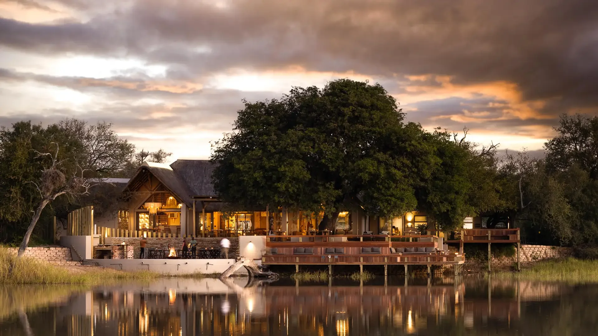 A lakeside resort scene during sunset with modern wooden building, outdoor fireplace, and large trees, reflecting in the water.