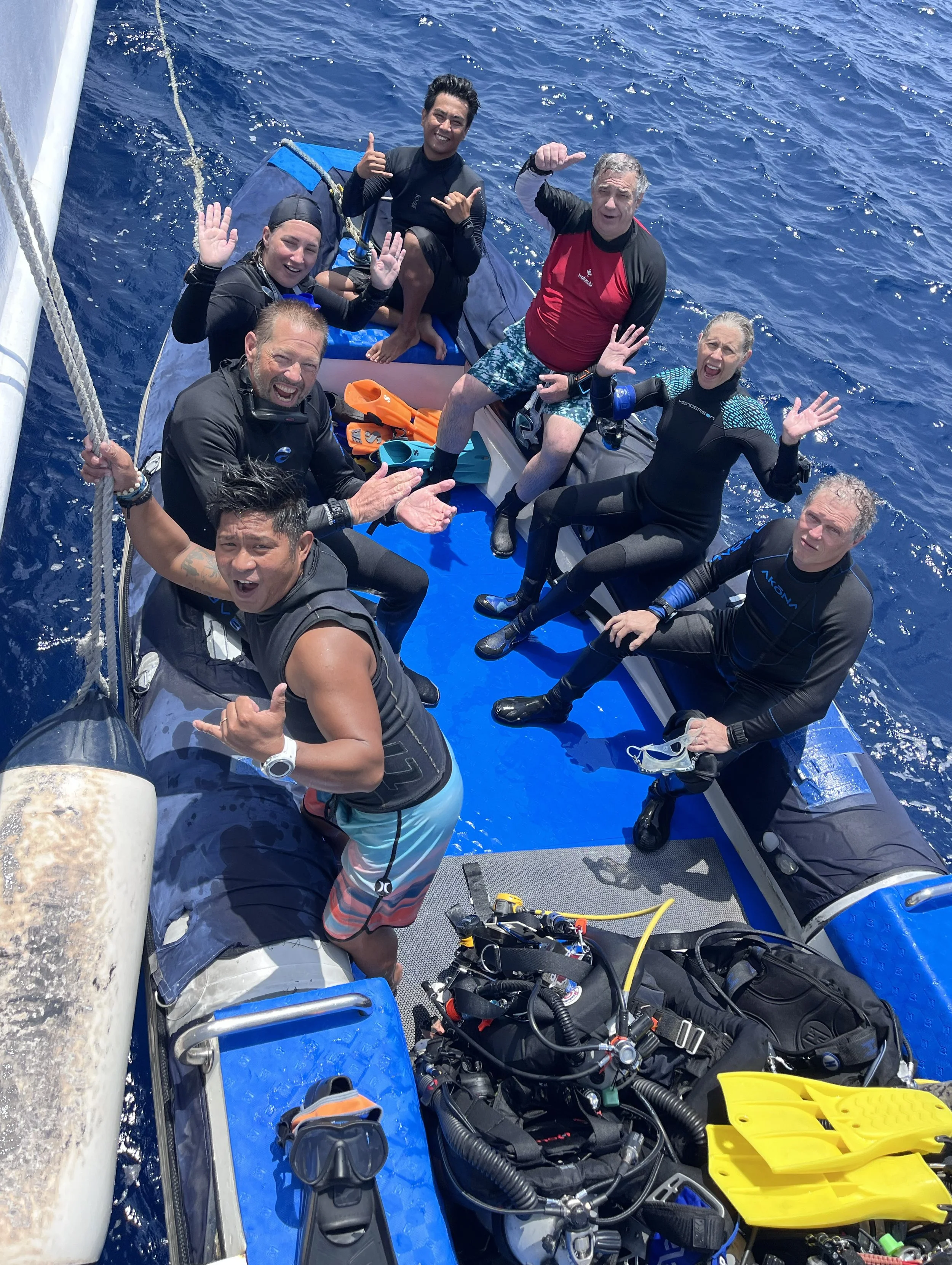 A group of six people on a boat, some wearing diving gear, celebrating with smiles and gestures, with deep blue ocean water surrounding them.