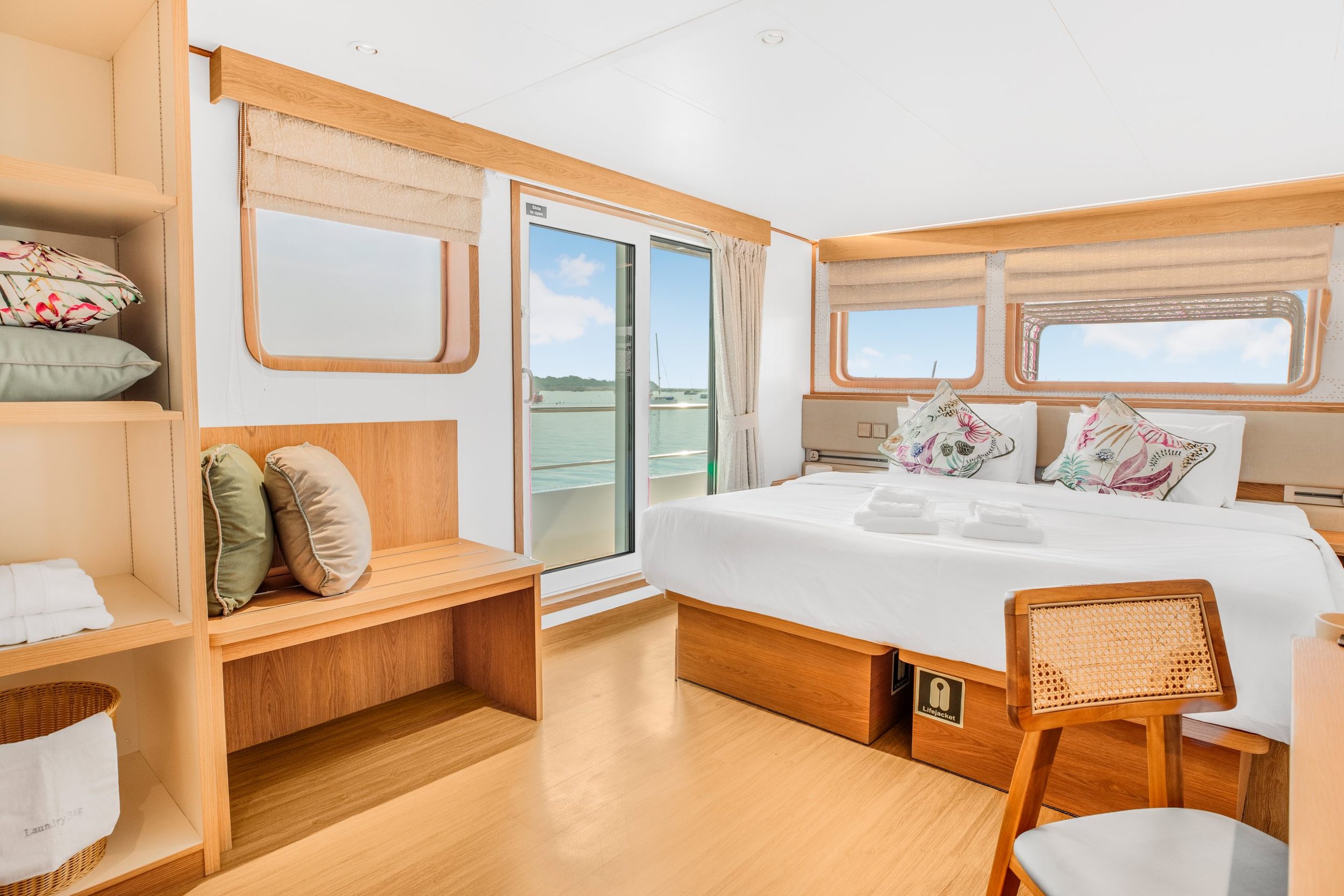 A bright, modern bedroom with large windows showing a view of the water and sailboats. The room has light wood furniture, a bed with decorative pillows, and a door leading to a balcony.