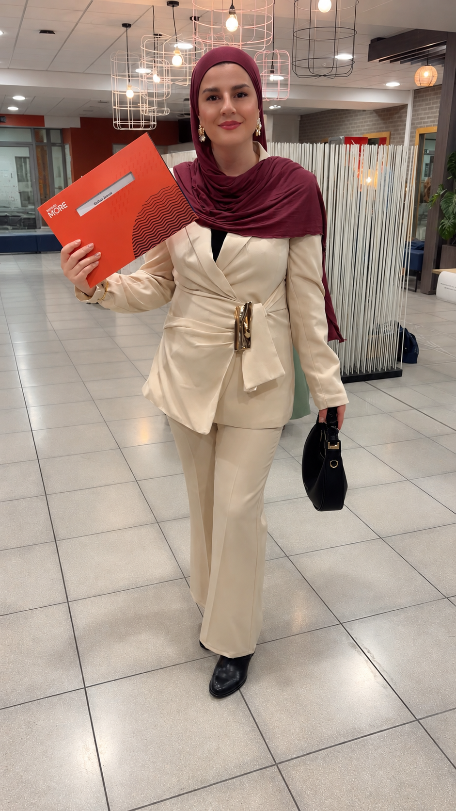 A woman dressed in a cream-colored suit with a maroon headscarf and earrings, holding a red folder and a black handbag, standing in a modern indoor space with geometric lighting fixtures.