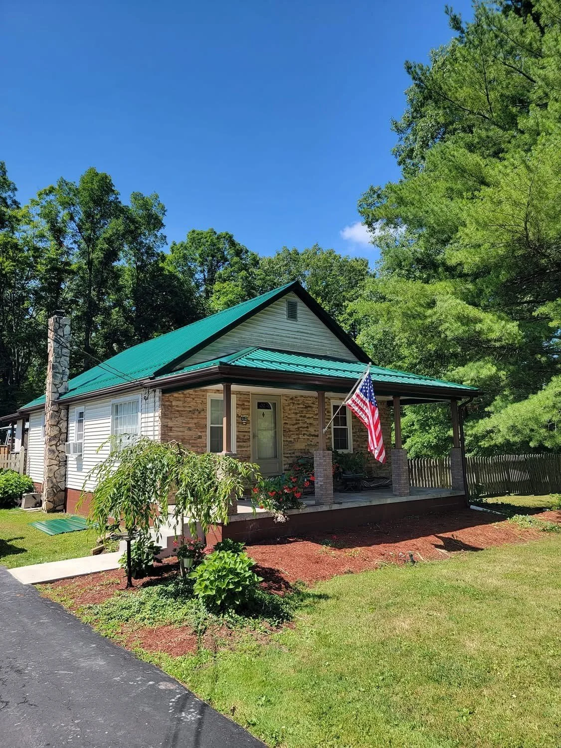 A house with a green metal roof and a front porch, decorated with an American flag, surrounded by trees and greenery under a blue sky.