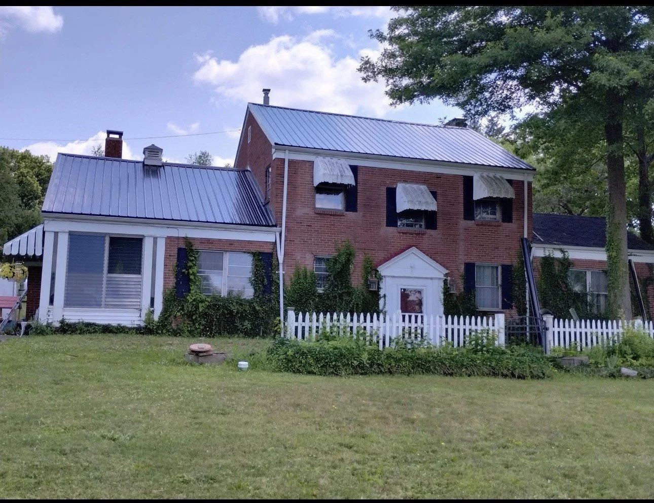 A two-story brick house with a white picket fence, small porch, and several windows with awnings, surrounded by greenery and tall trees, under a partly cloudy sky.