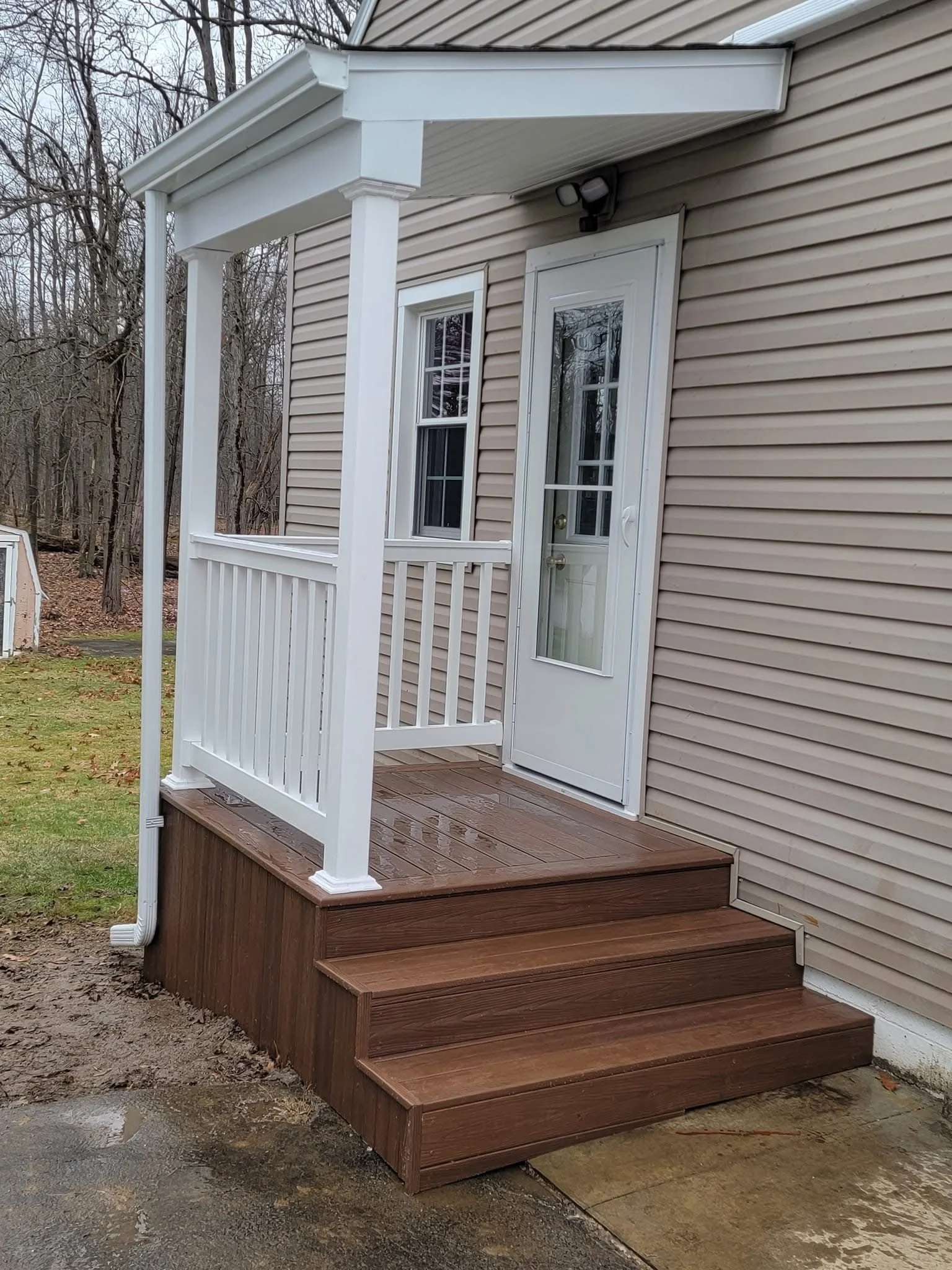 A small wooden deck with stairs leading up to a back door on a beige house. The deck has a white railing and a white porch with a metal roof. The ground is muddy with patches of grass.