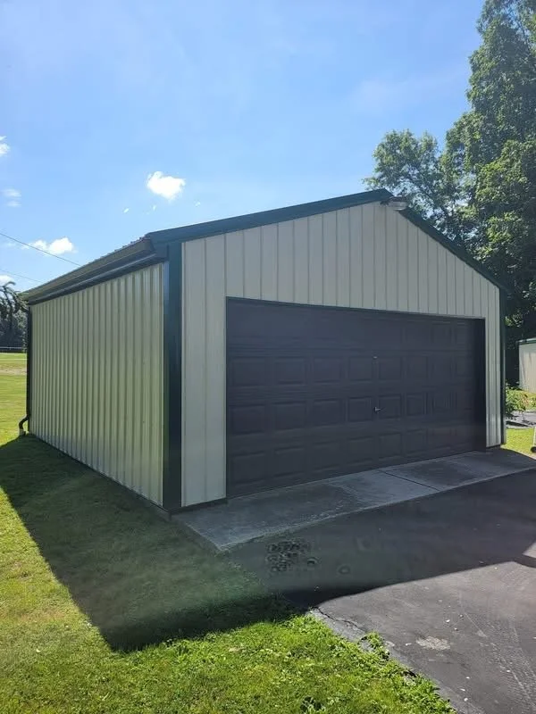 A metal garage with beige siding and a dark brown roll-up door, located on a paved driveway with a grassy yard, under a clear blue sky with some clouds.