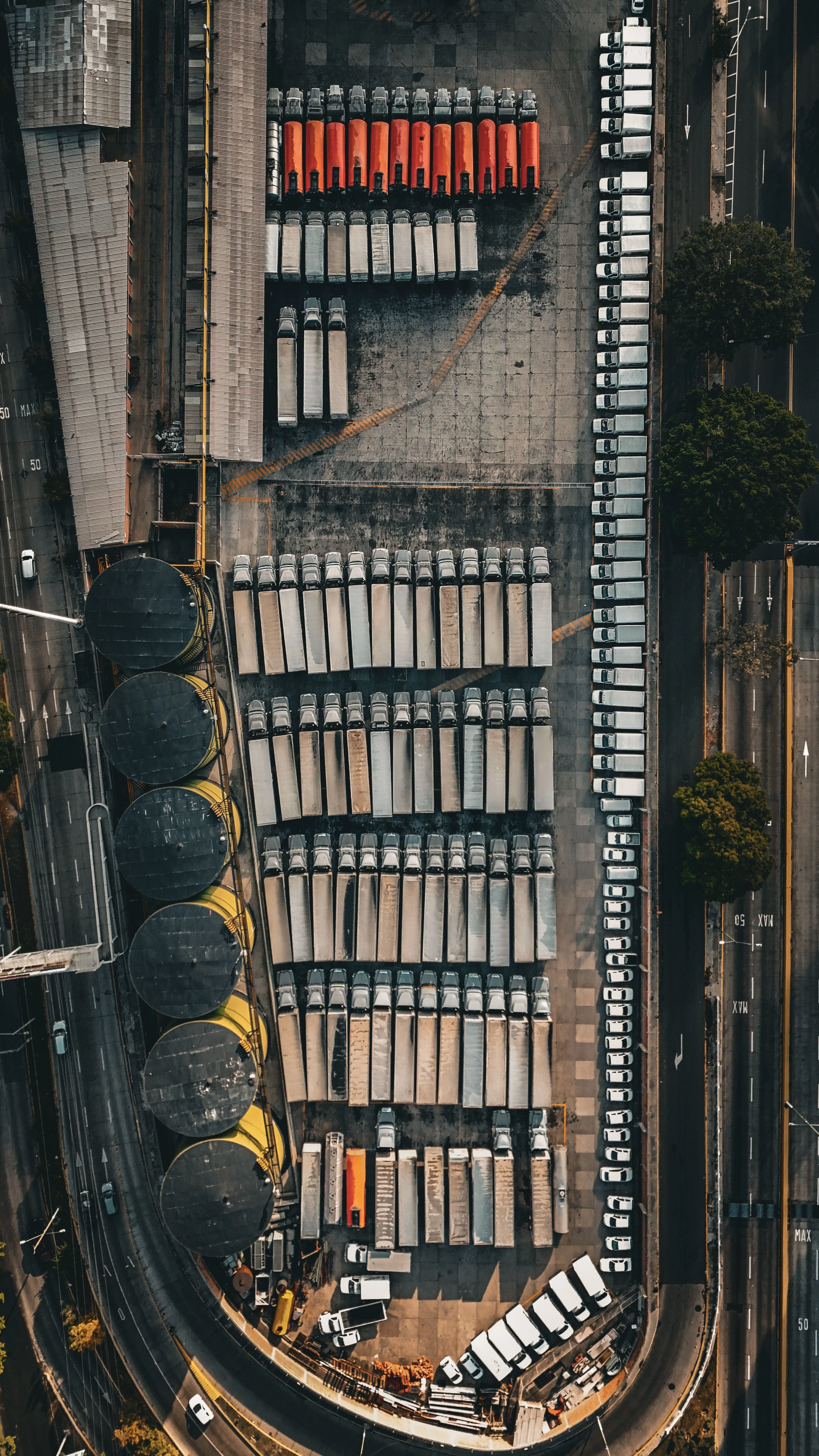 Aerial view of a parking lot with rows of trucks, cars, and buses, surrounded by roads and trees.