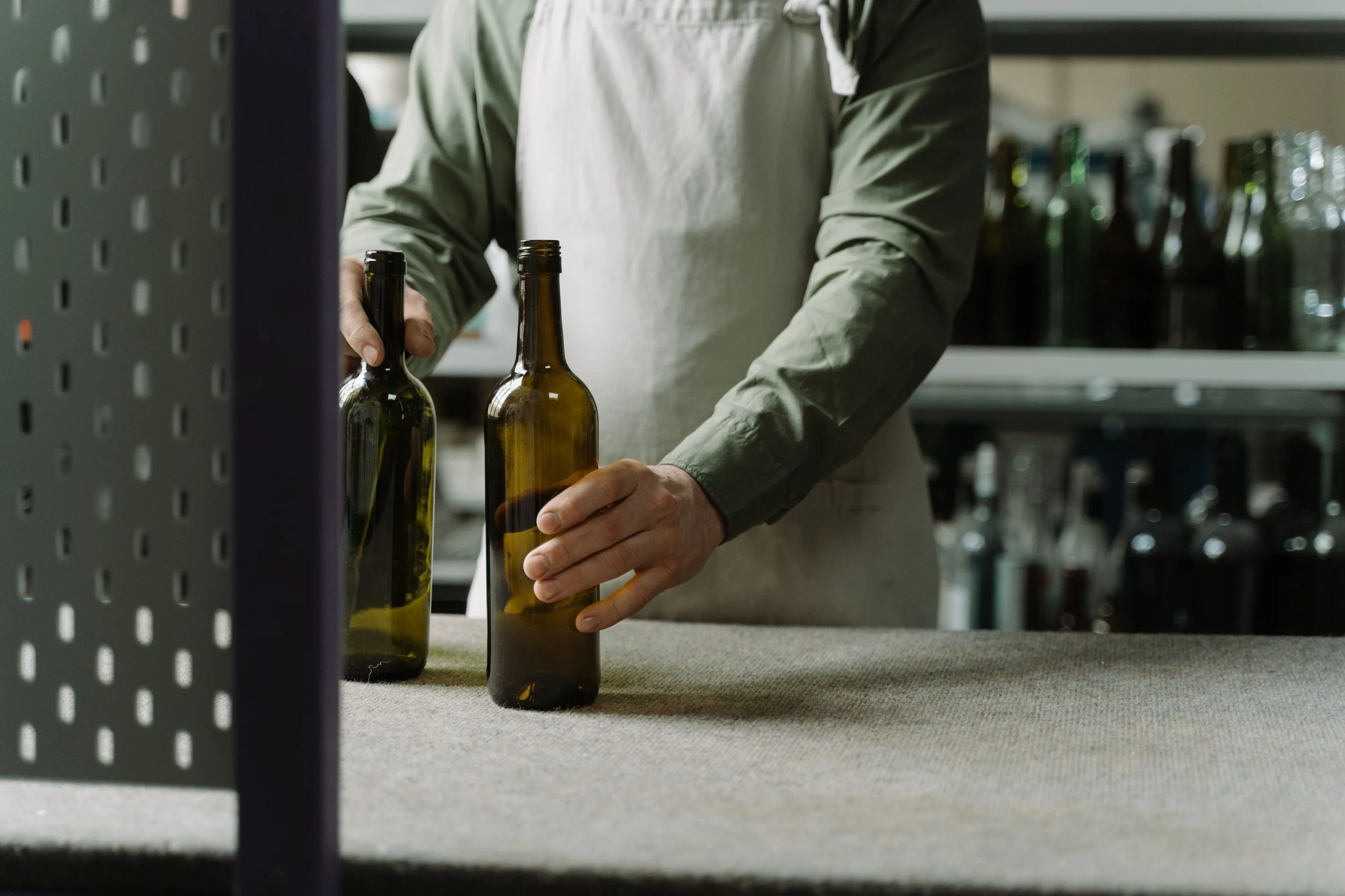 Person in a green shirt and white apron placing beer bottles on a counter in a store or bar.