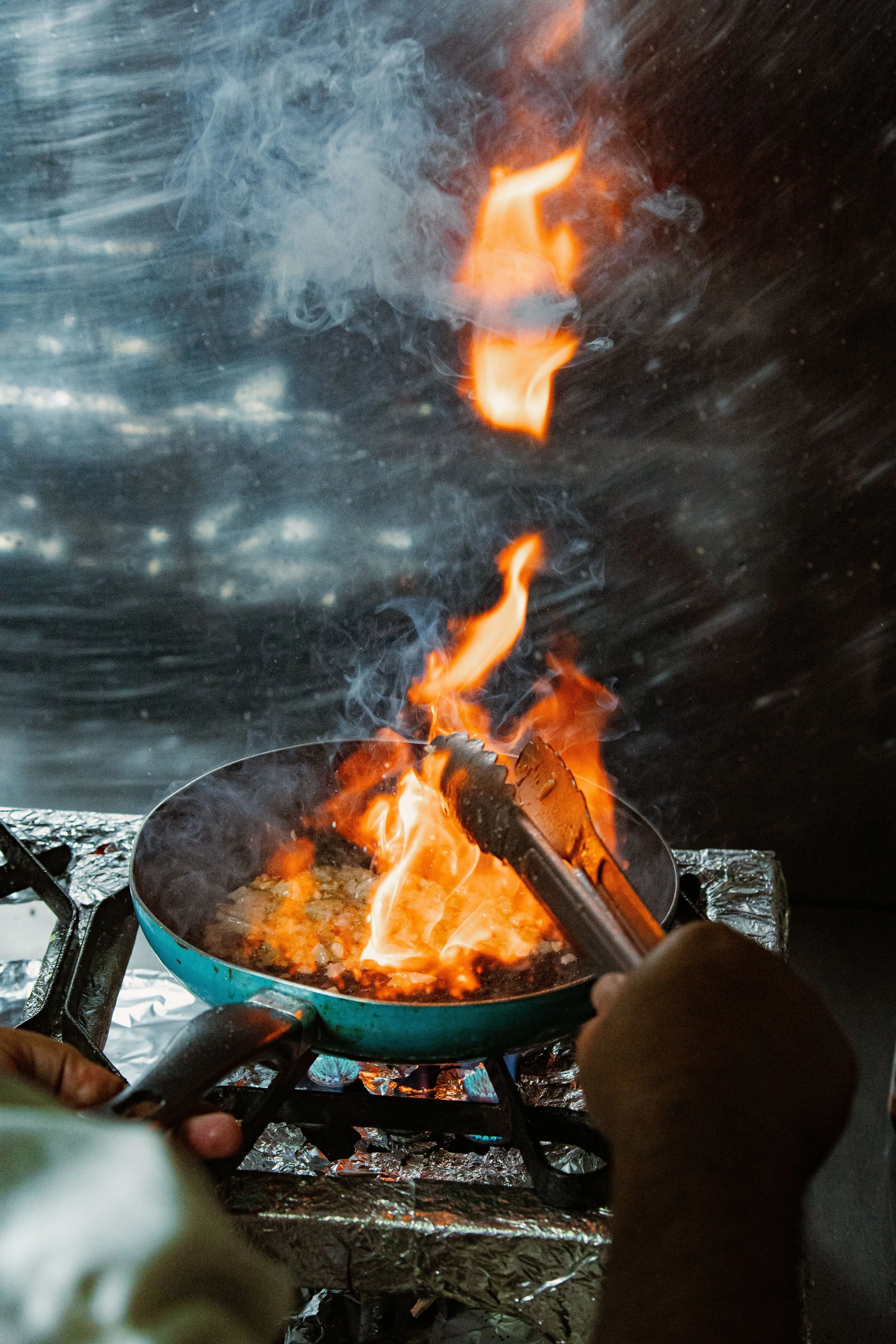 A person is cooking food over an open flame on a stove, with flames rising from a frying pan, in a setting with aluminum foil-covered surfaces.