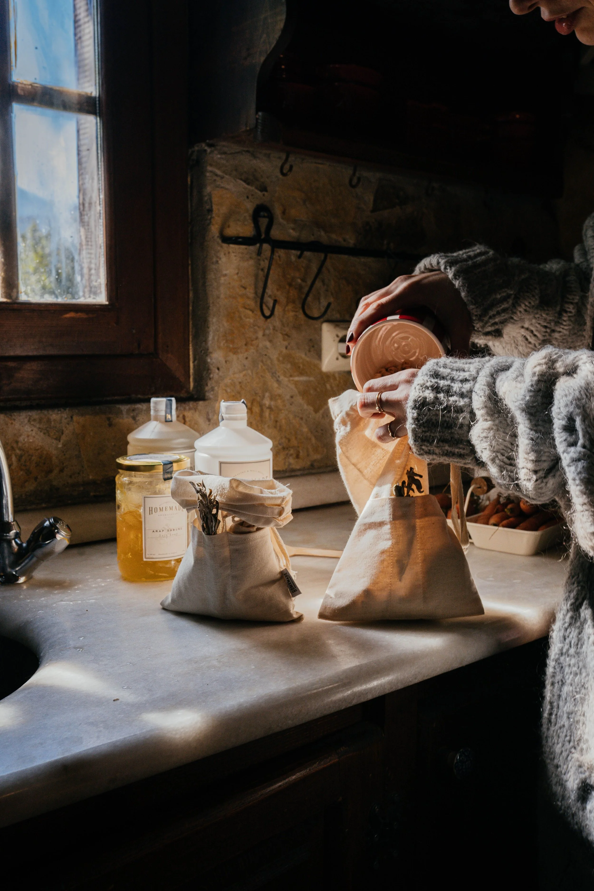 A person wearing a gray knitted sweater packing cookies into paper bags in a rustic kitchen with stone walls, wooden window frame, and various jars and containers on the counter.