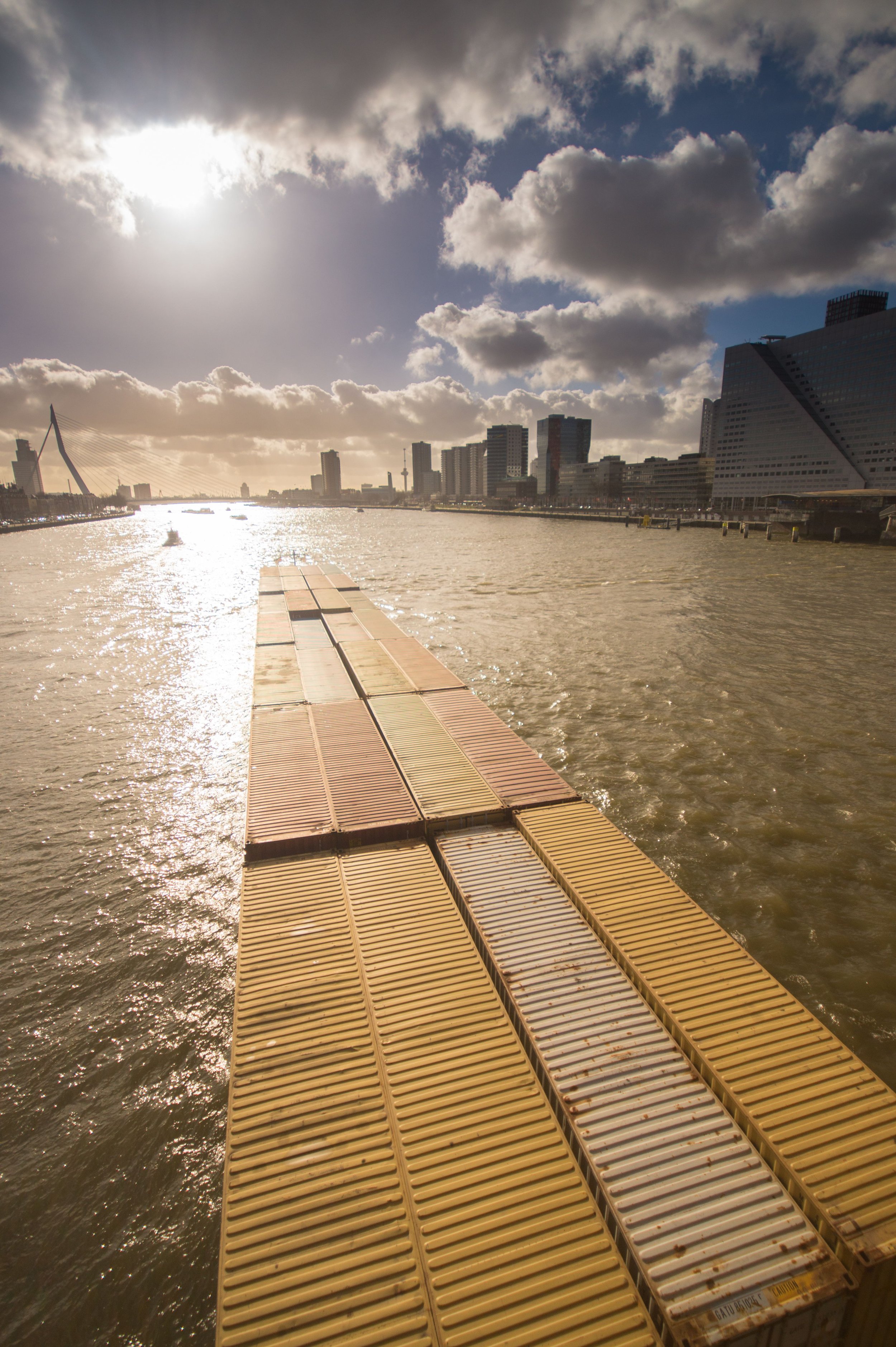 A dock made of yellow and gray floating platforms extends into a river in a city, with buildings, a bridge, and a partly cloudy sky in the background.