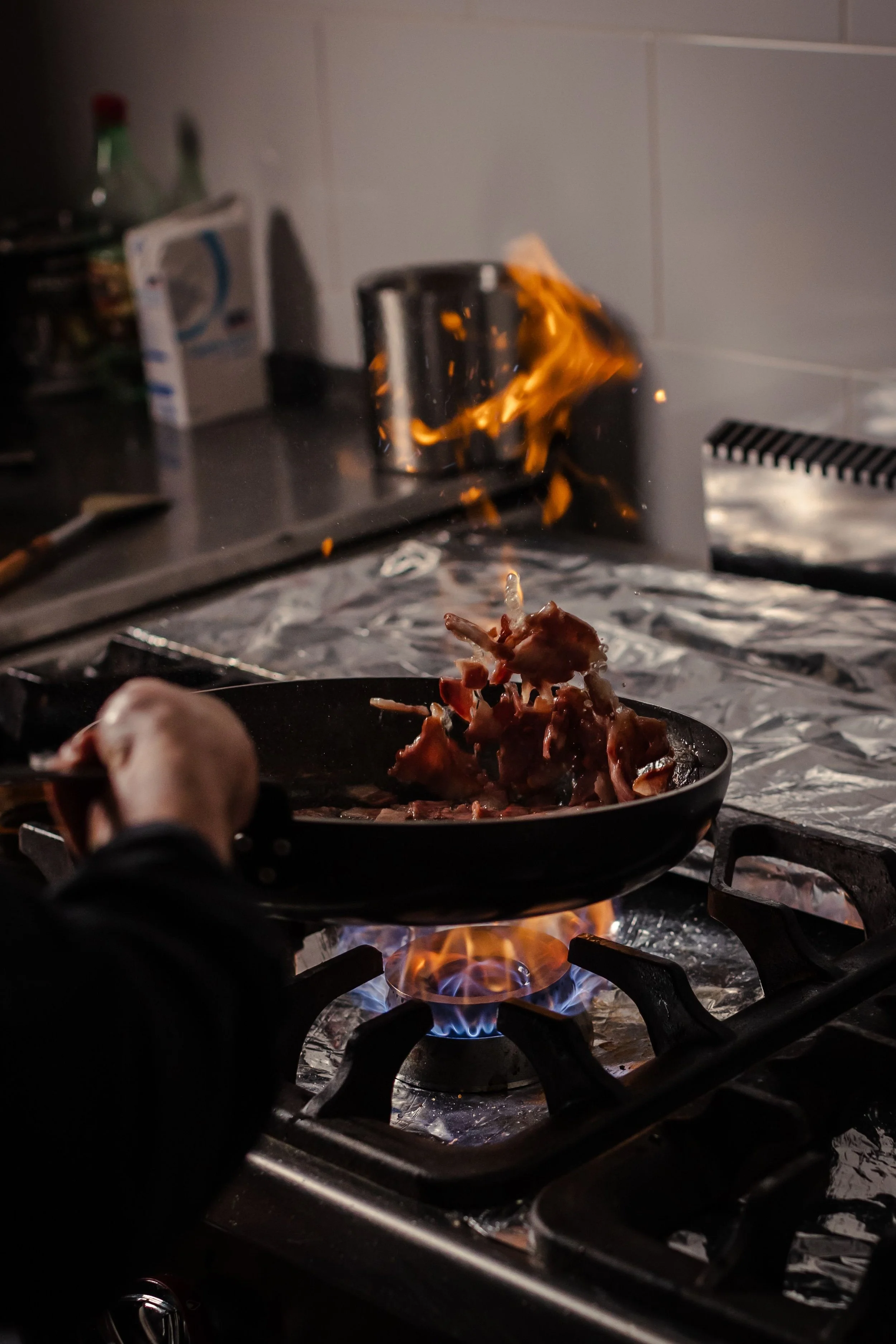 A person cooks meat in a frying pan over an open flame on a kitchen stove. Flames rise around the pan, and kitchen items are visible in the background.