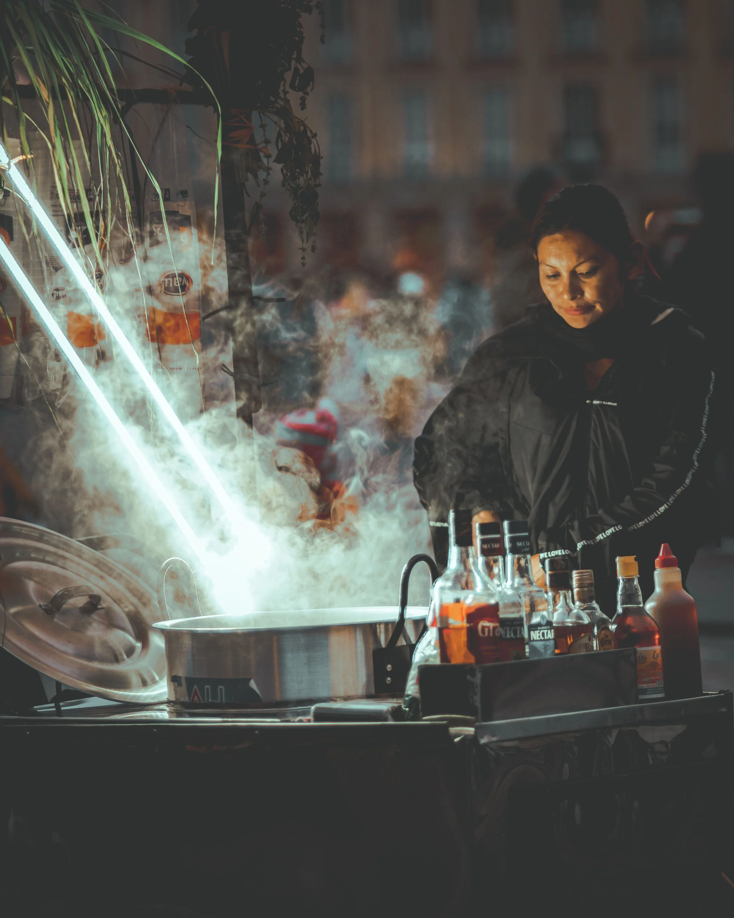 A woman in a dark jacket standing behind a food cart with bottles of condiments on it, in an urban environment with a building in the background and smoke or steam rising from the cart.