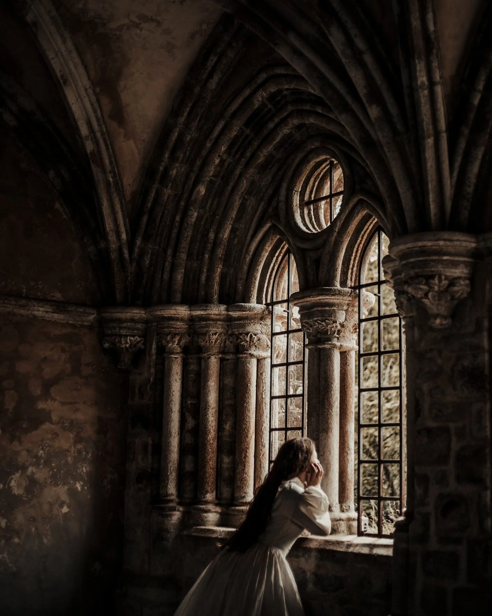 A woman in a white dress sitting inside a gothic cathedral, looking out of a large window with intricate stone columns and archways, holding her head in her hands.