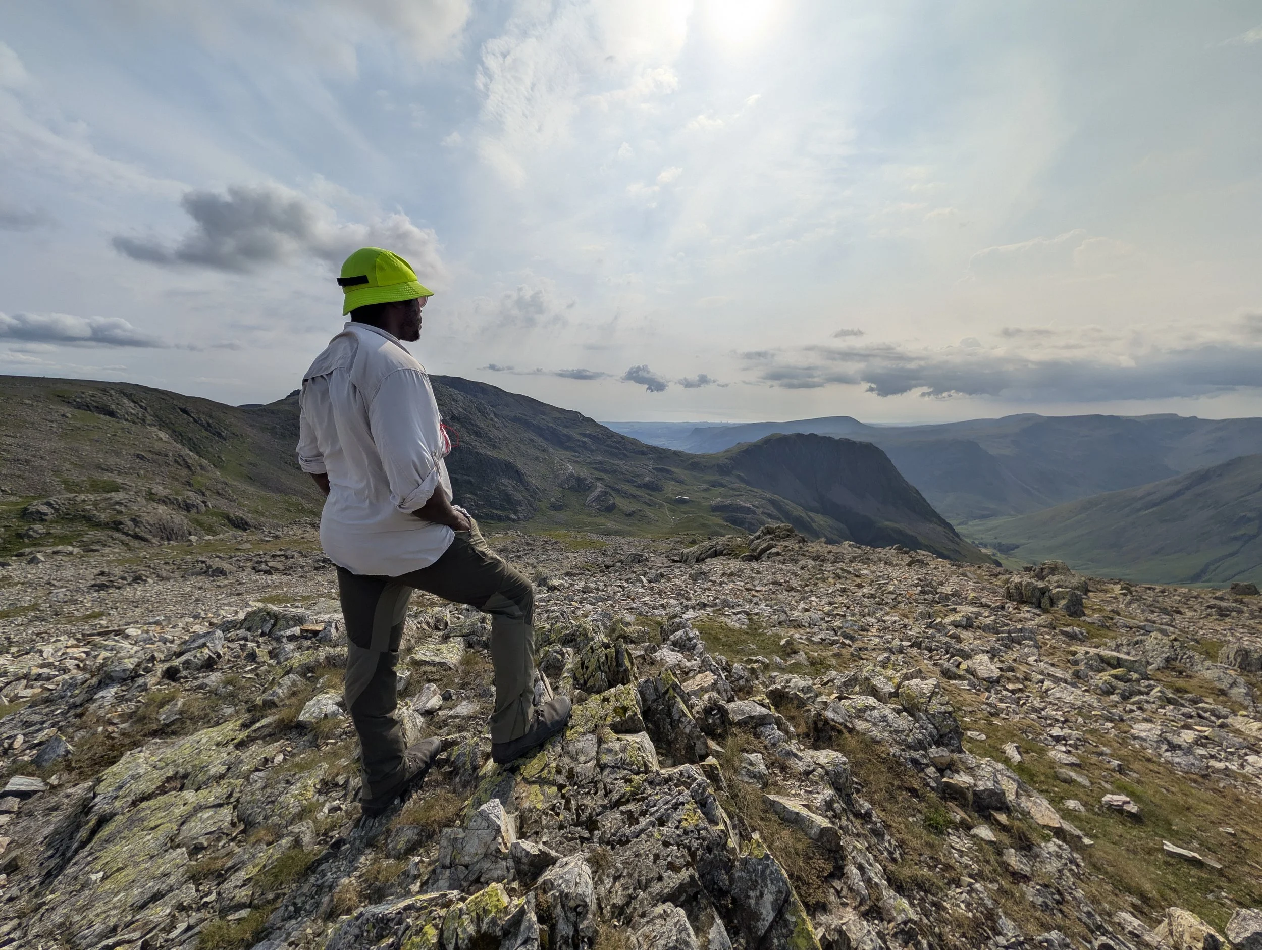 A person standing on rocky terrain overlooking a valley with mountains in the distance under a partly cloudy sky.