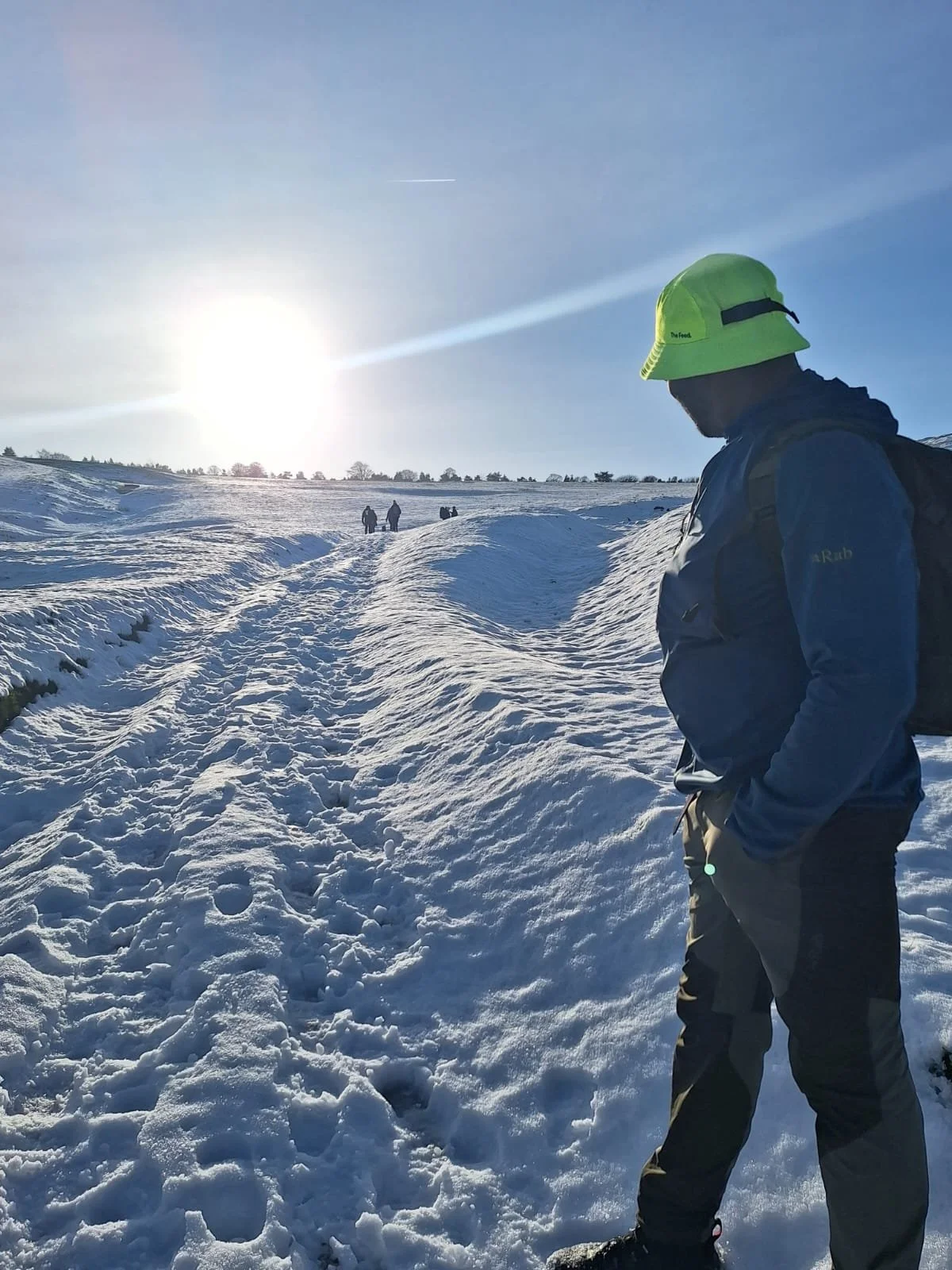 A person in winter clothing, including a yellow hat and dark jacket, standing in a snow-covered landscape under a bright sun, with others walking in the distance.