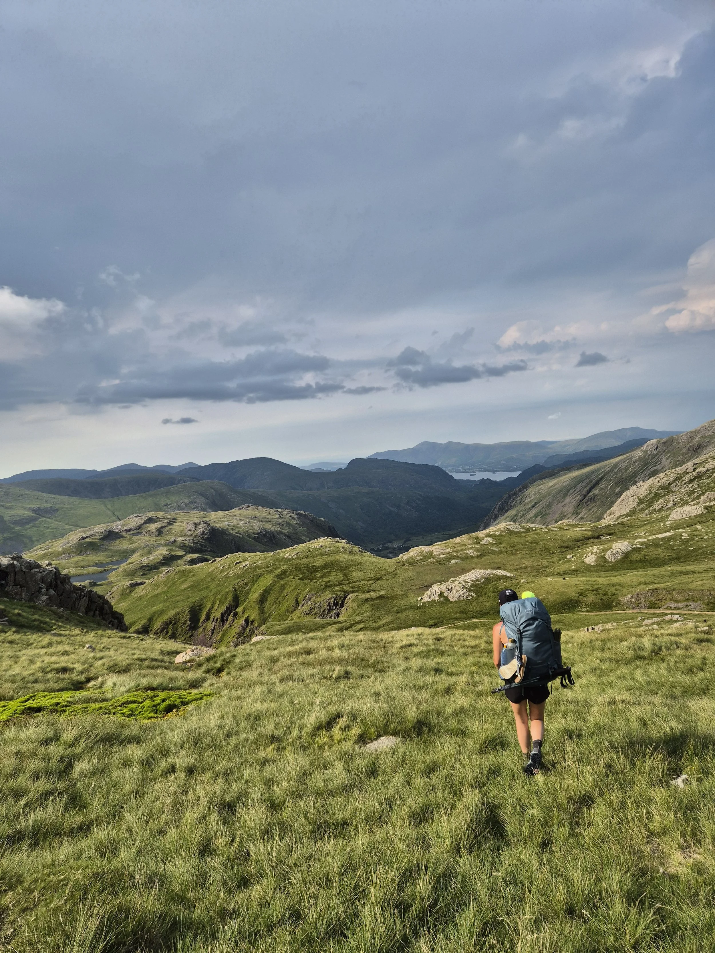 A hiker walking through a lush green mountain landscape with rolling hills, distant mountains, and cloudy sky in the background.