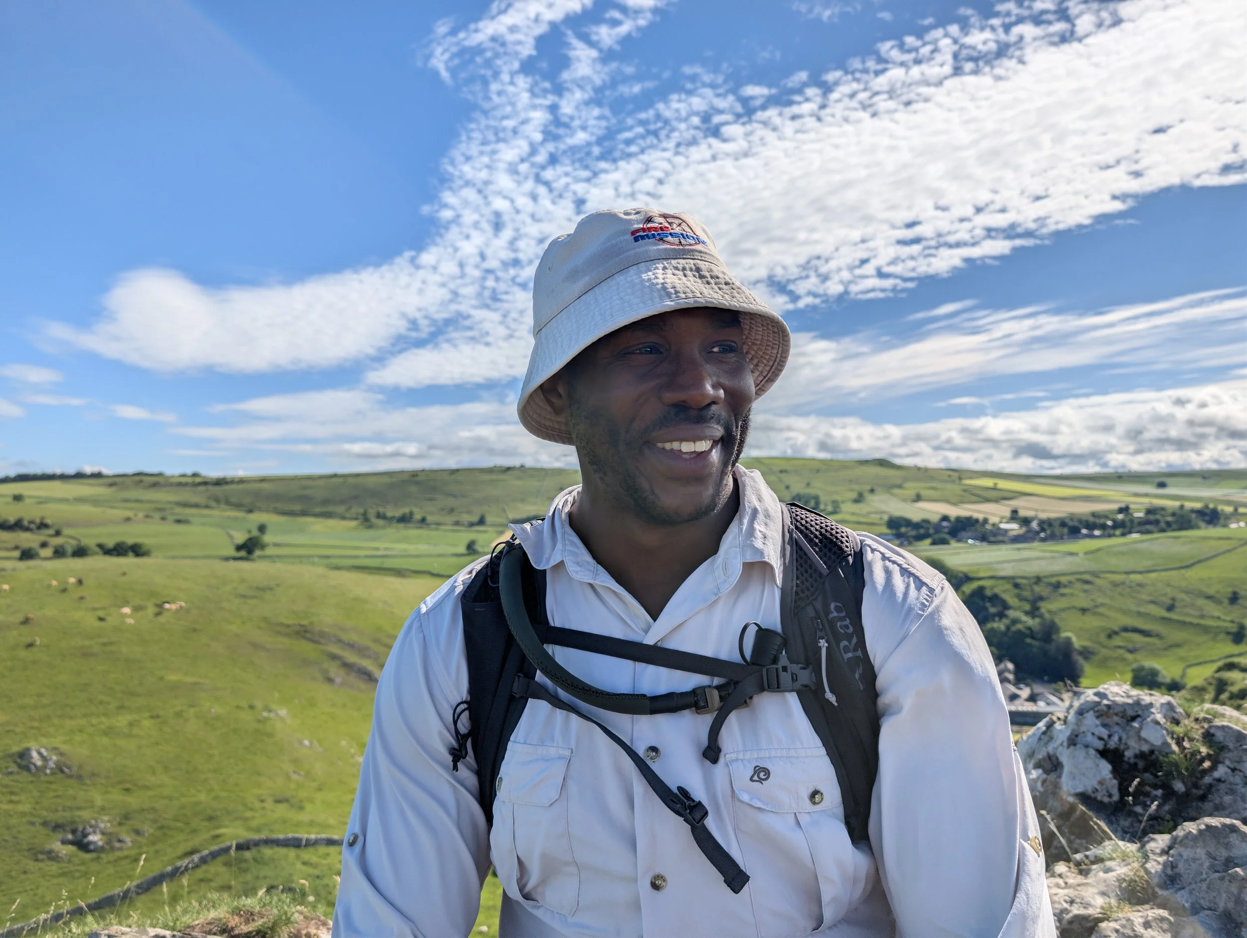 A man wearing a beige bucket hat with a logo, a white long-sleeve shirt, and a black backpack standing outdoors in a green landscape with a blue sky and clouds.