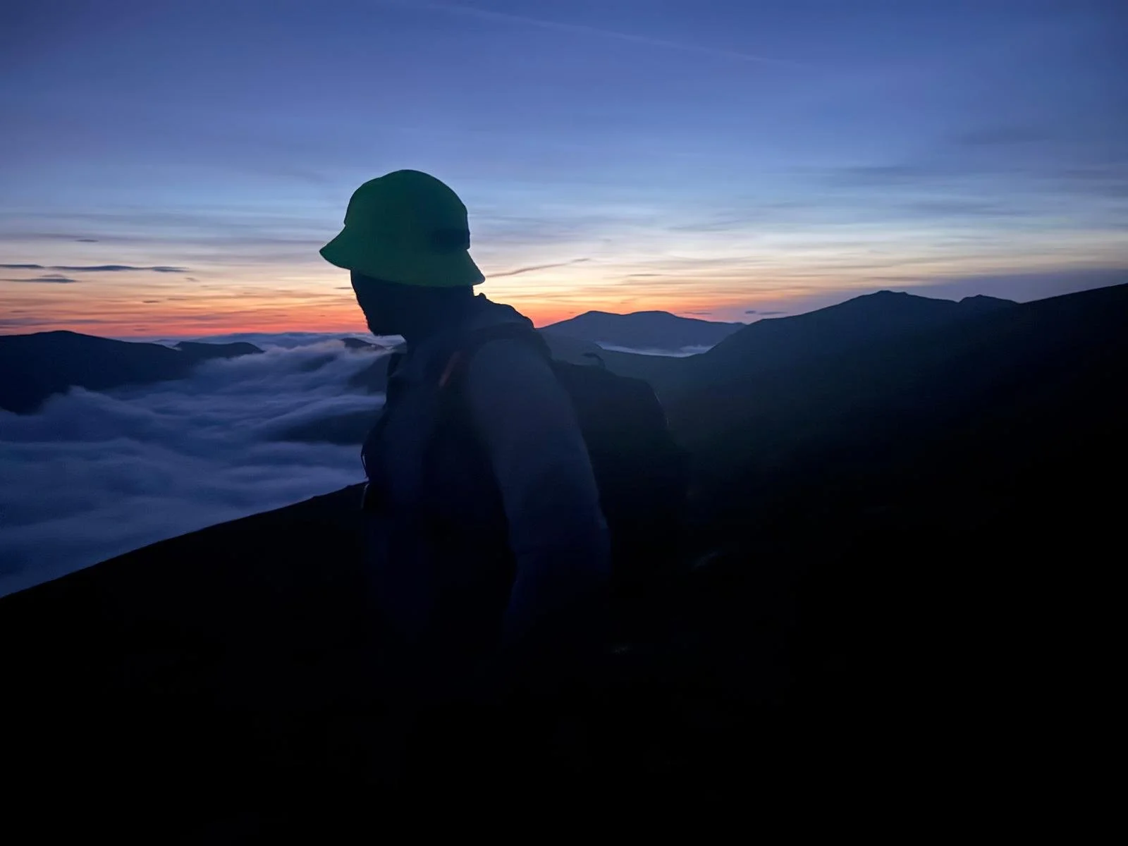Silhouette of a person wearing a hat and backpack standing on a mountain at sunset, with clouds below and mountains in the distance.