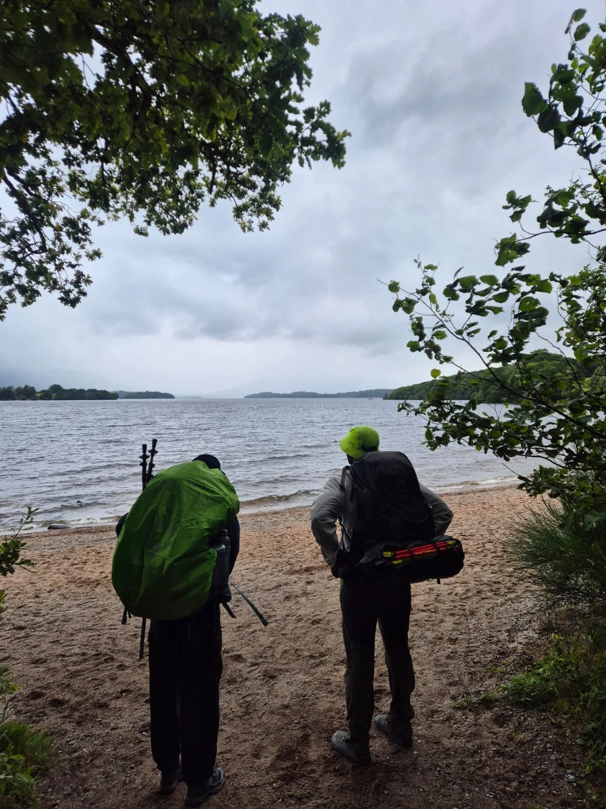 Two people with backpacks and outdoor gear standing on a sandy beach, looking out over a large body of water under a cloudy sky, surrounded by trees.