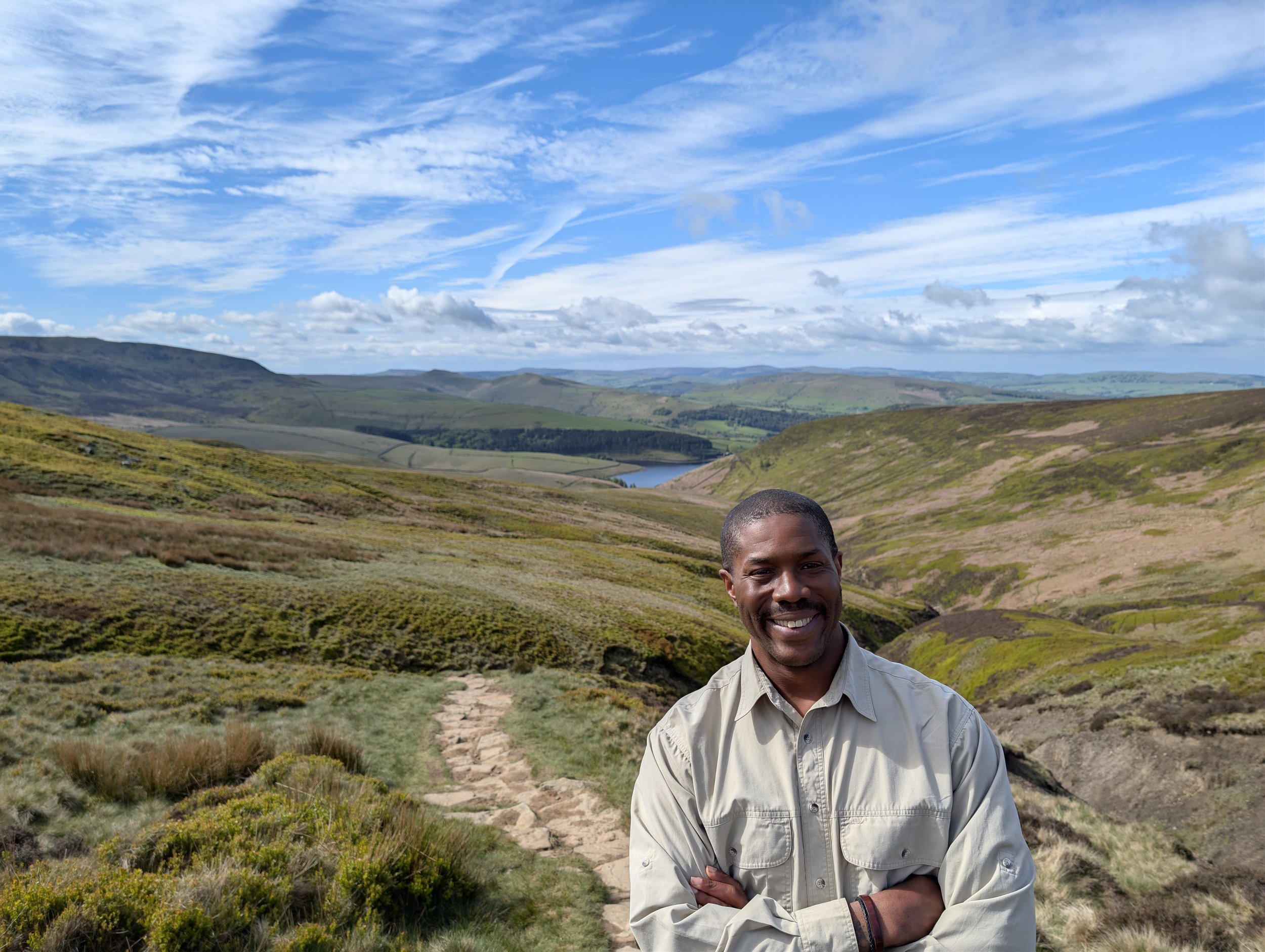 Smiling man wearing a beige jacket standing on a trail in a scenic hilly landscape with green fields, a lake, and blue sky with clouds.