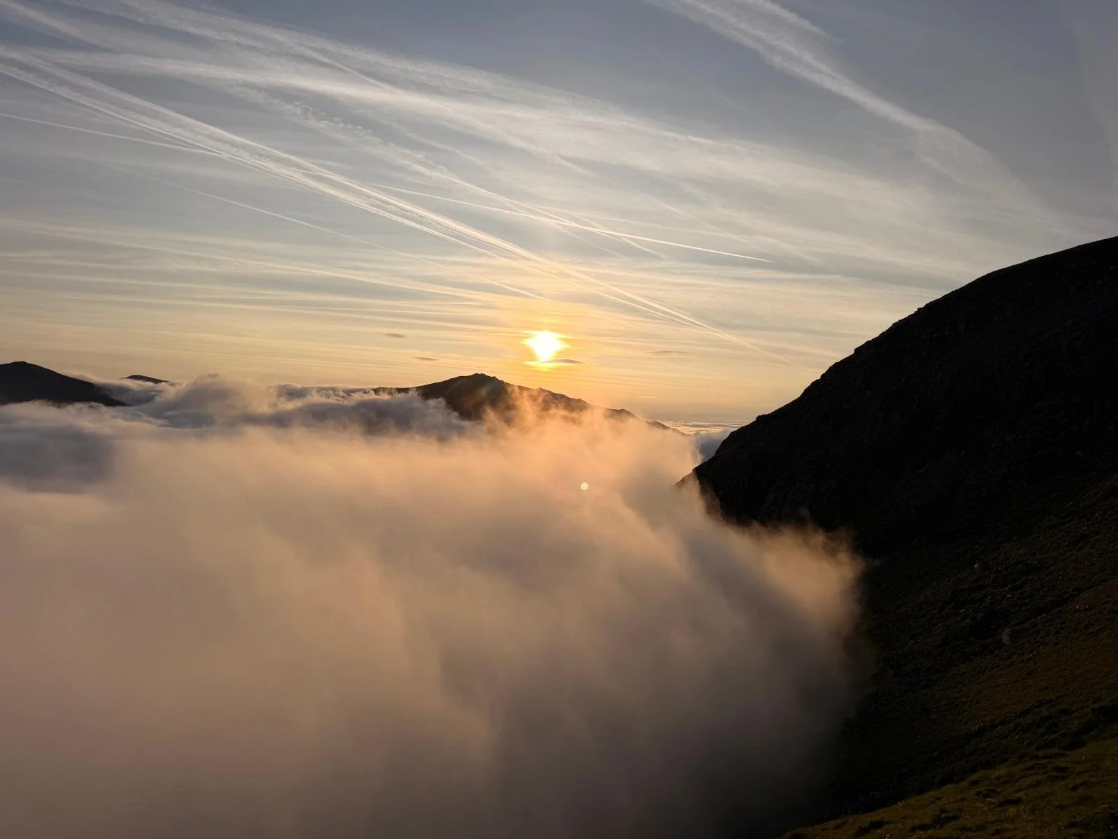 Mountain landscape at sunrise with clouds below, vapor rising from the terrain, and contrails streaking across the sky.