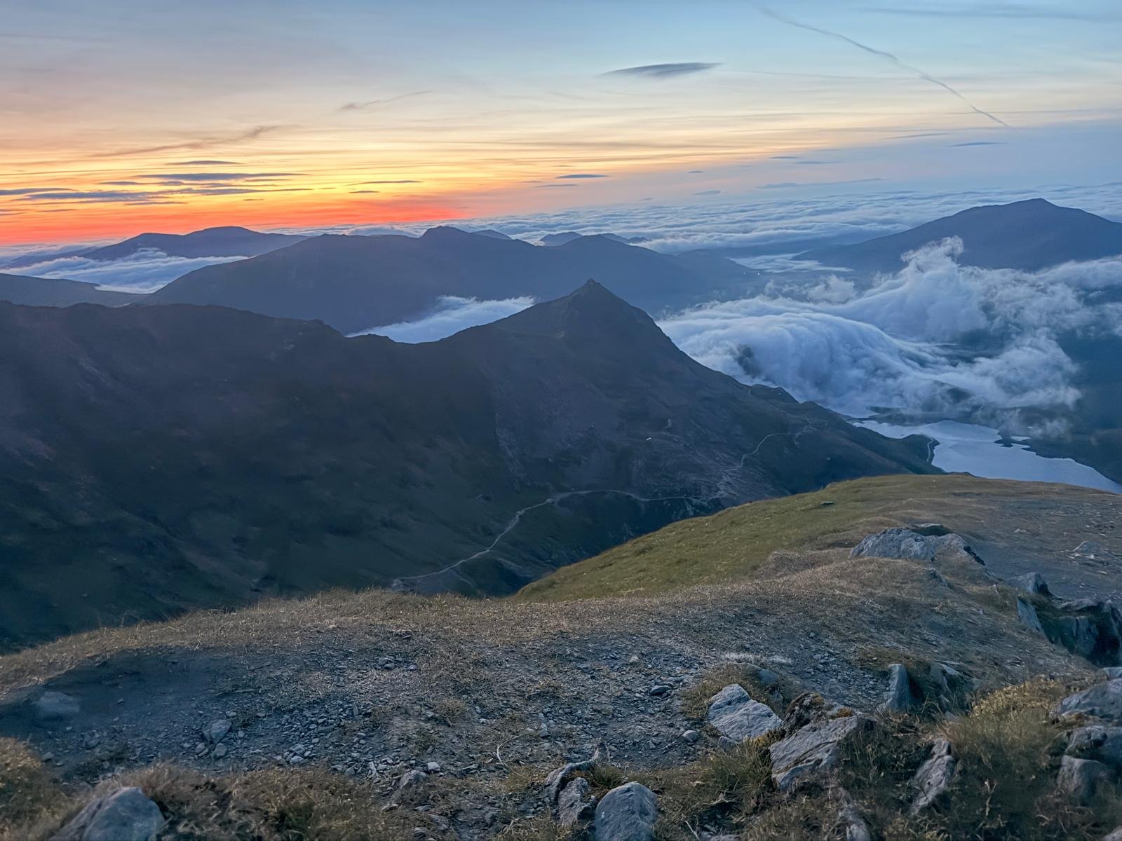 A mountain landscape at sunrise with a sky of orange and blue hues, cloud layers among mountain peaks, and a grassy slope with rocks in the foreground.
