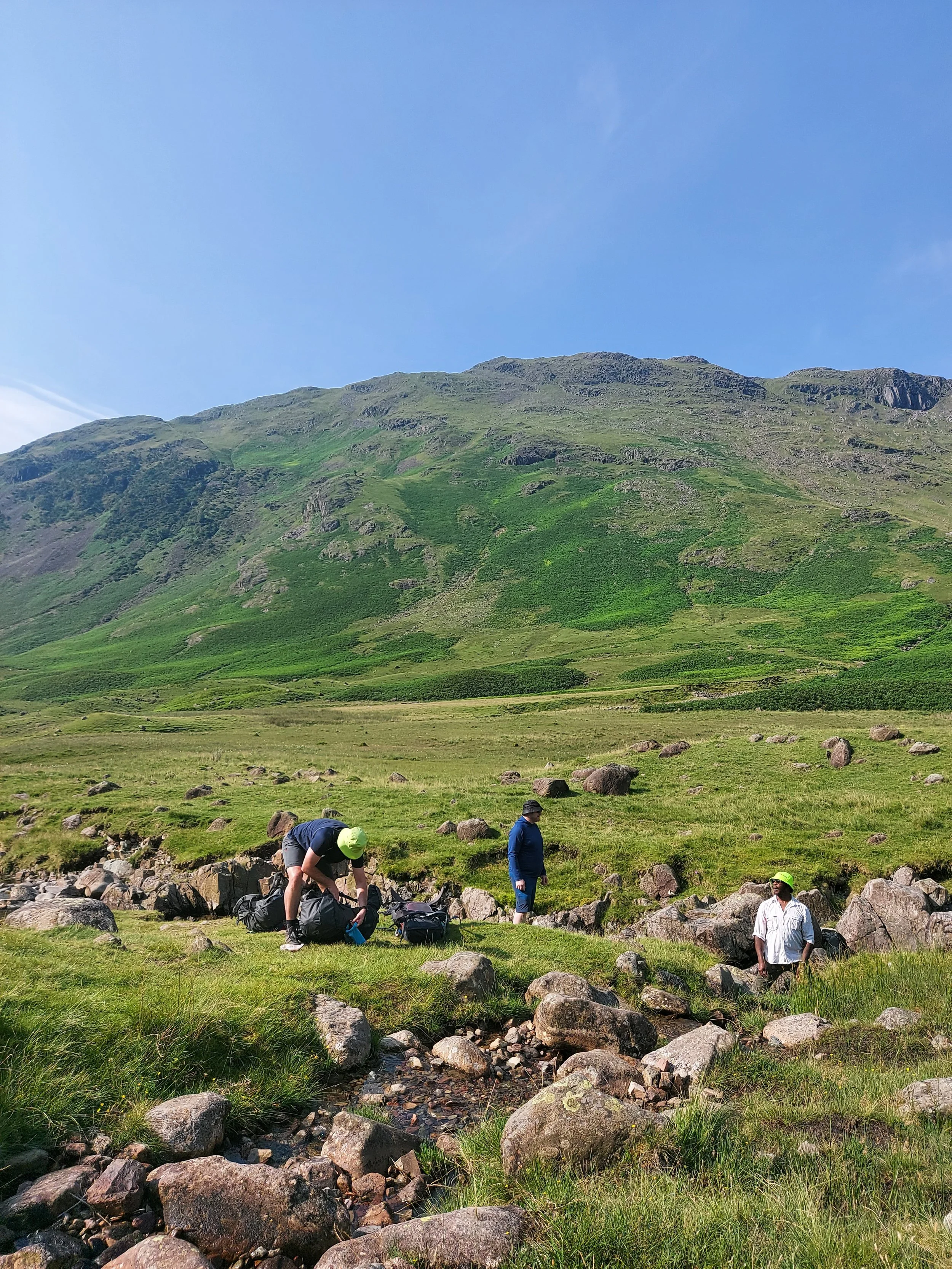 Three people in a grassy, rocky area near a stream with a mountain in the background. One person is bending down in front, another standing in the middle, and the third standing to the right, all dressed in outdoor gear.
