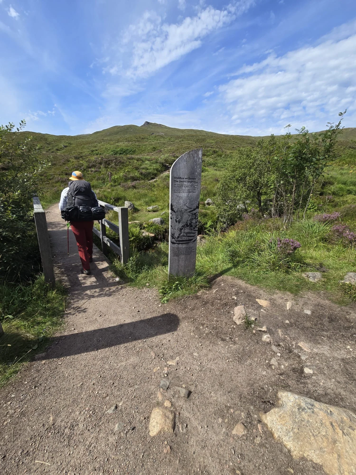 A hiker with a backpack and hat walking across a small wooden bridge in a grassy landscape with a hill in the background. There is a large gray stone with black writing next to the path, and trees and wildflowers surrounding the area. The sky is partly cloudy with blue sky visible.